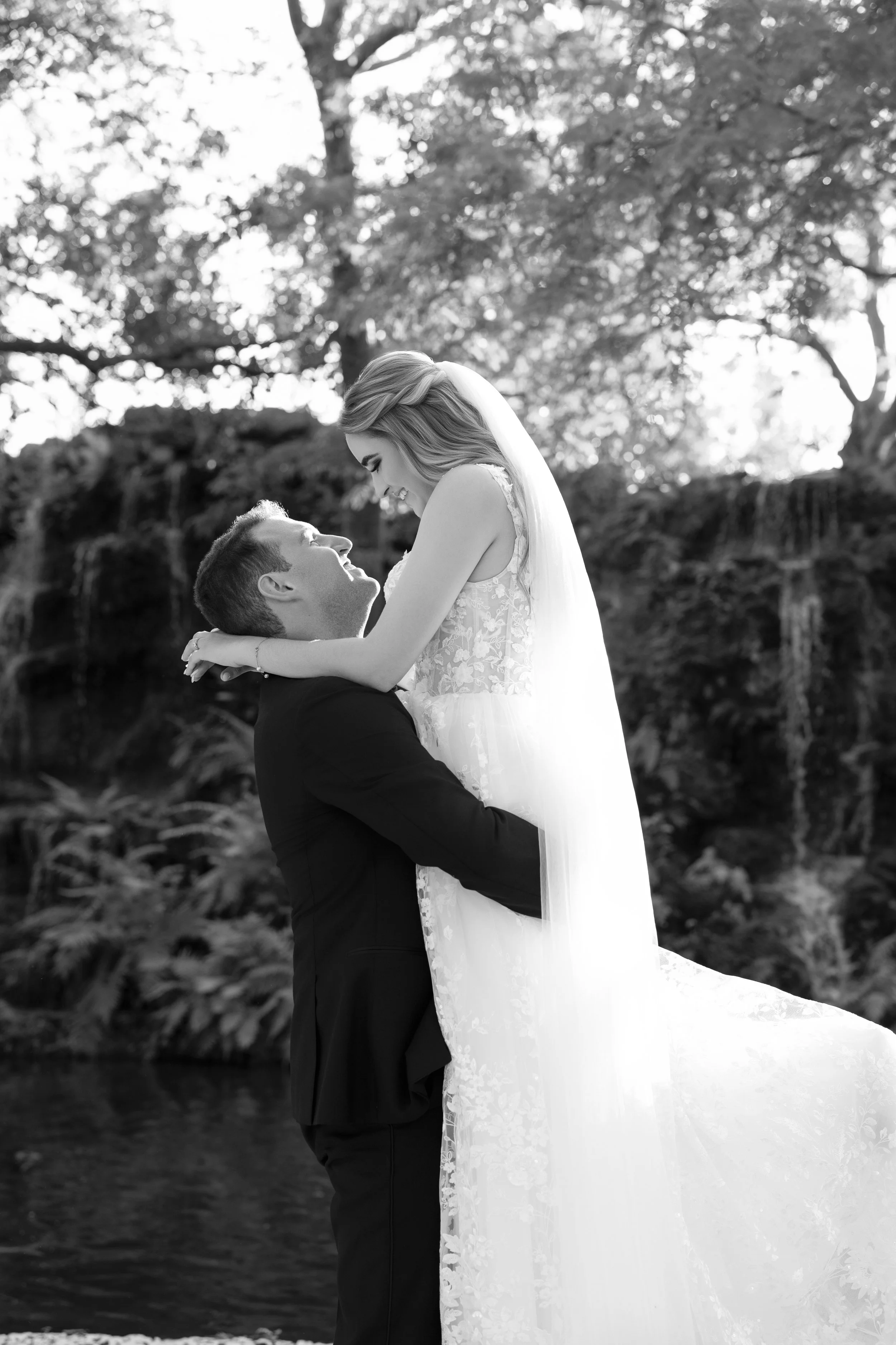 A wedding couple in formal attire, with the groom holding the bride in a loving embrace outdoors, surrounded by trees and a waterfall, in black and white.