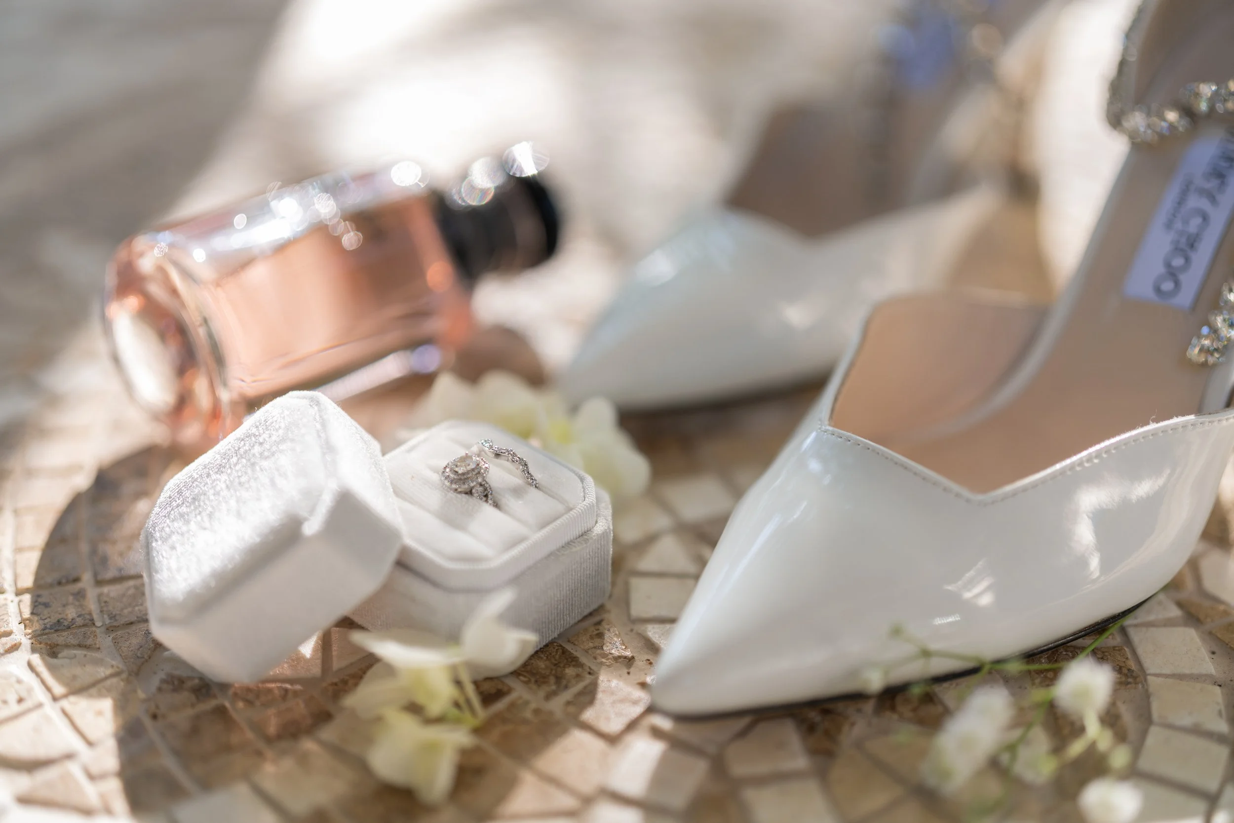 A pair of white wedding shoes, a jewelry box with a diamond ring, a bottle of perfume, and small white flowers on a textured surface.