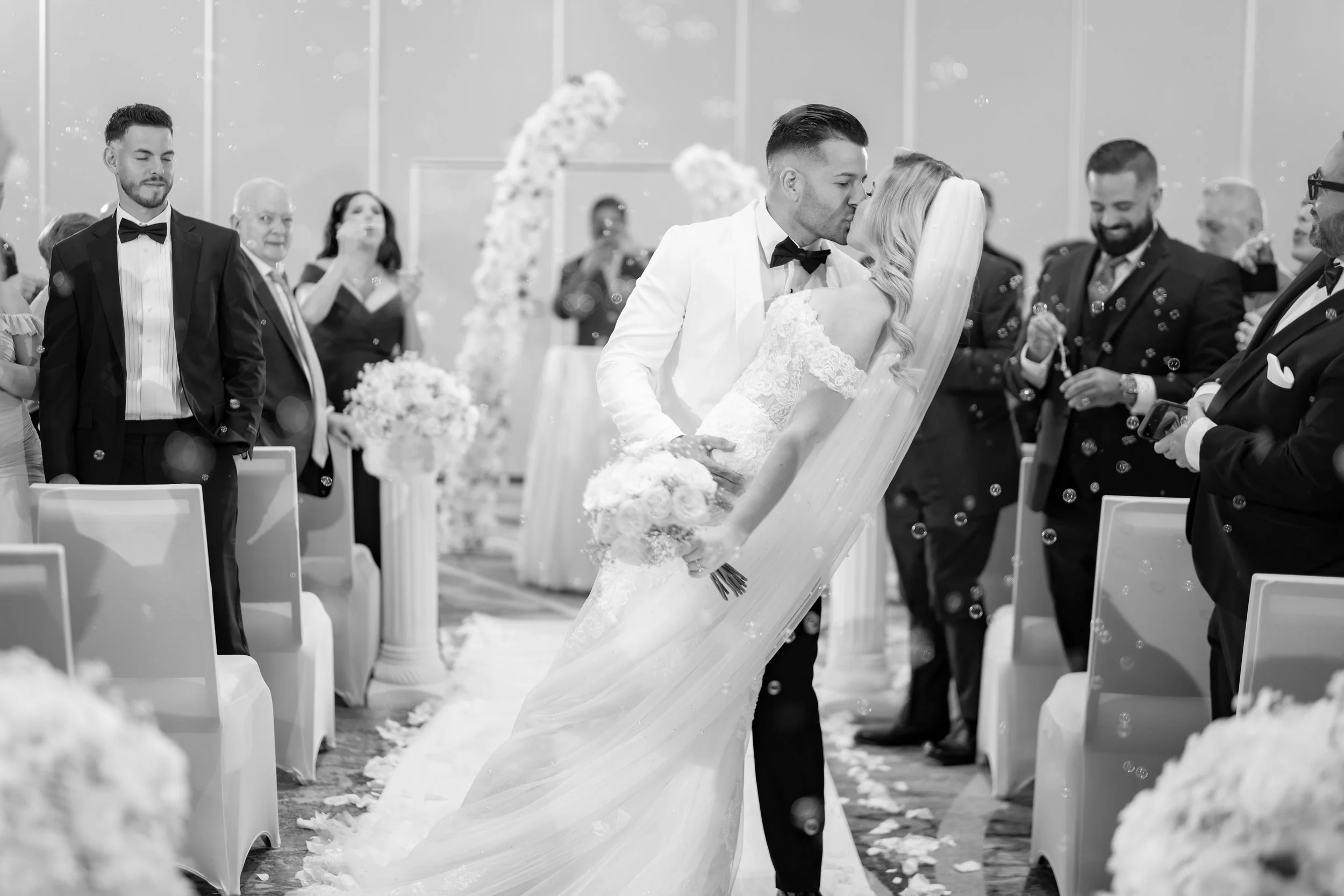 A black and white photo of a wedding ceremony, showing a couple kissing while standing on a wedding aisle decorated with flowers. Guests are standing and clapping on both sides, with some taking photos.