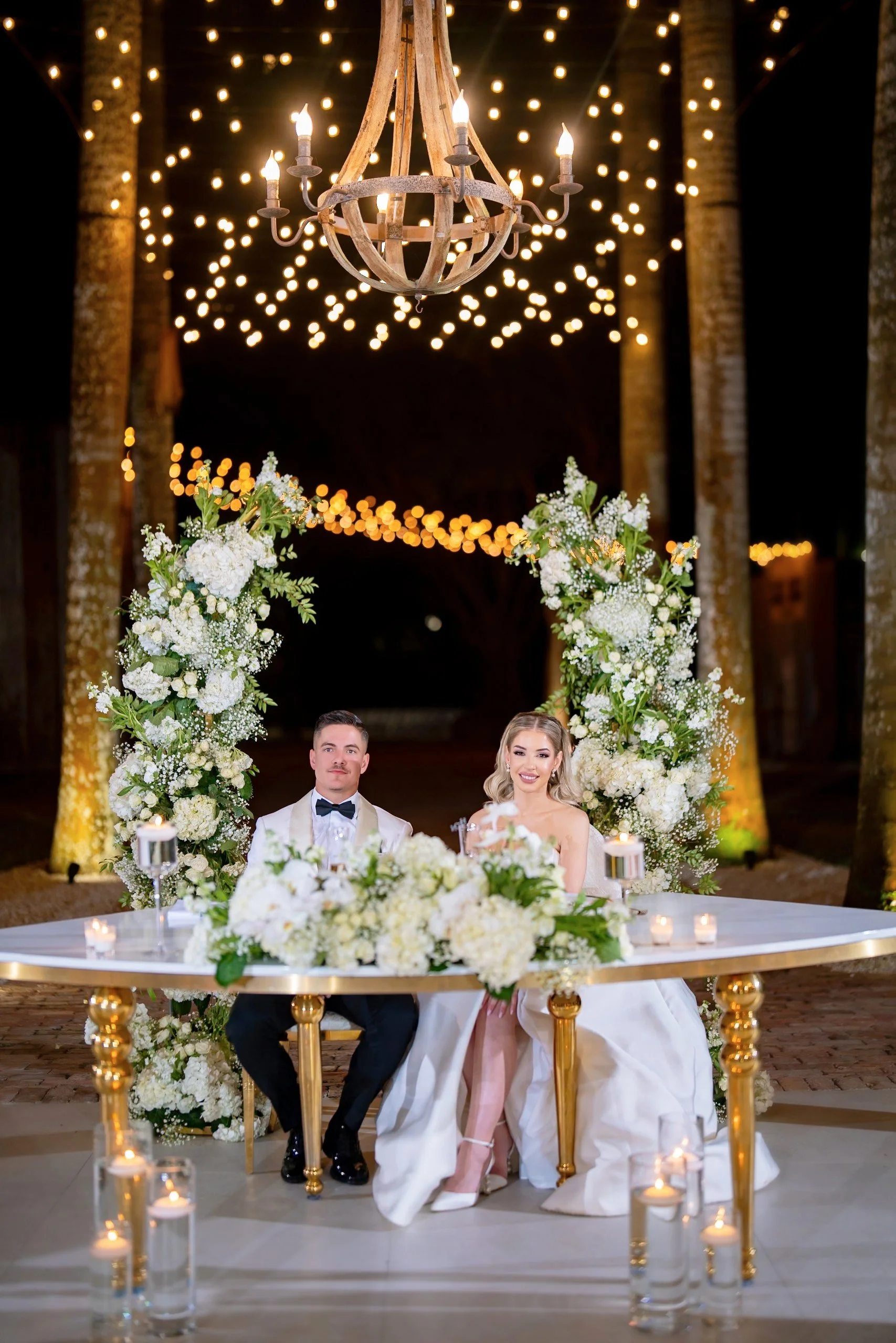 A bride and groom sitting at a decorated wedding reception table outdoors at night, with hanging string lights and floral arrangements.