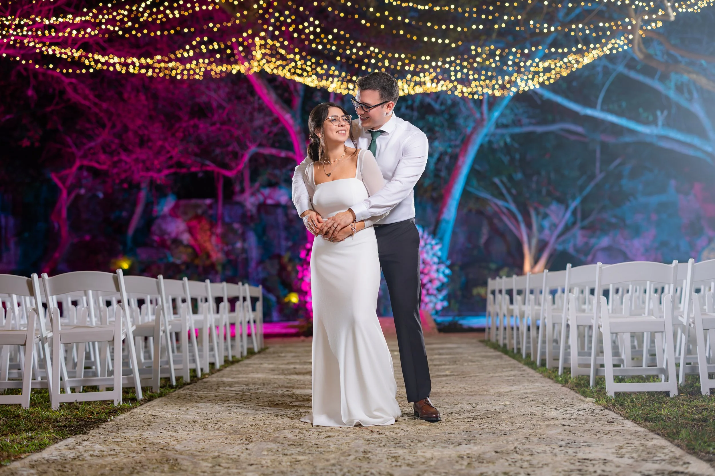 A couple in wedding attire dancing on a pathway outdoors at night, surrounded by white chairs, under string lights with colorful trees in the background.