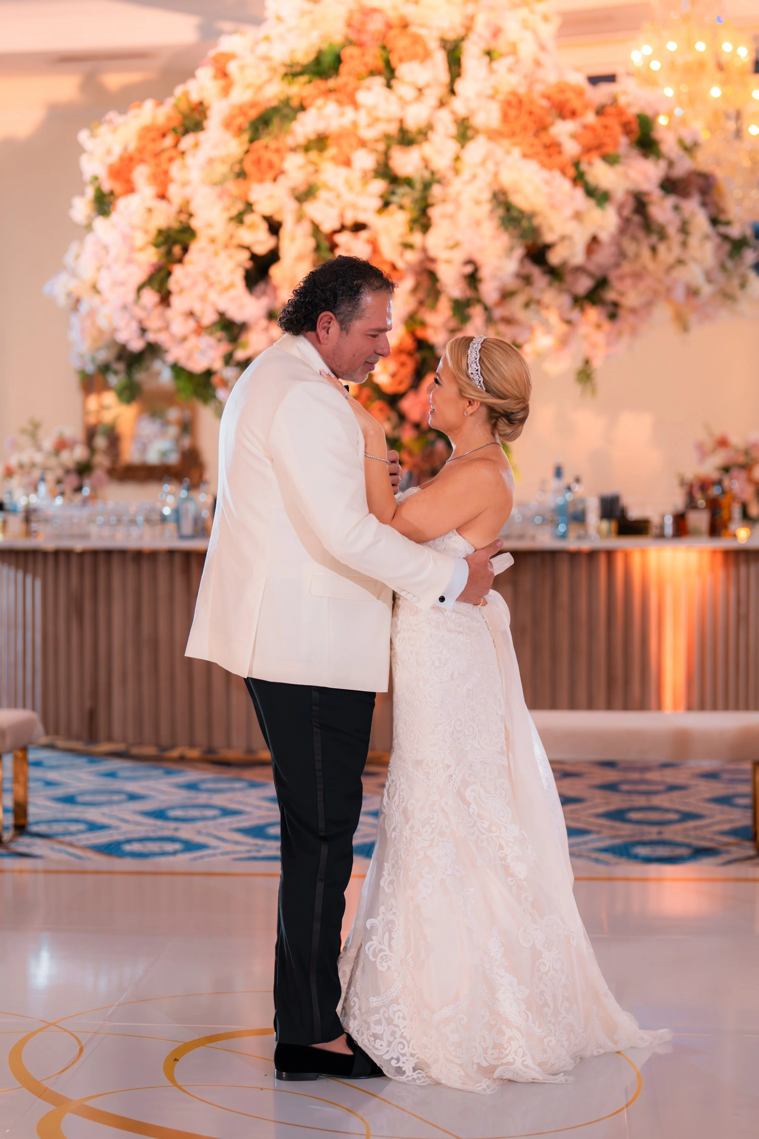 A bride and groom dance at their wedding reception, with a large floral arrangement and a decorated bar in the background.