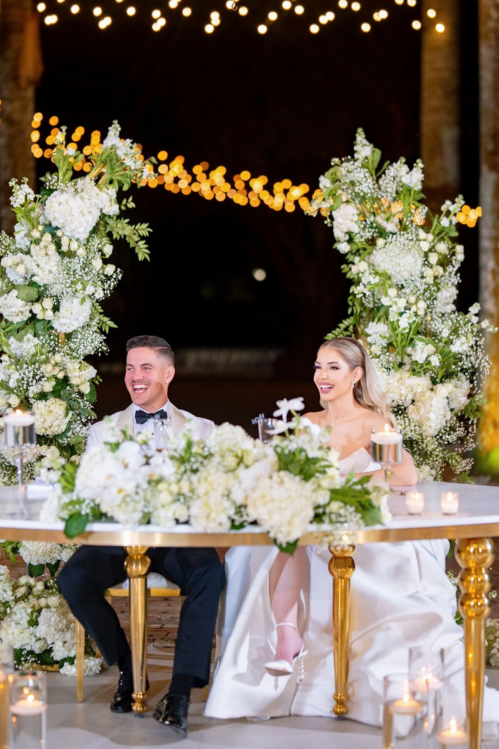 A bride and groom sitting at a wedding reception table decorated with white flowers and surrounded by floral arrangements and candles, under a canopy of string lights at night.