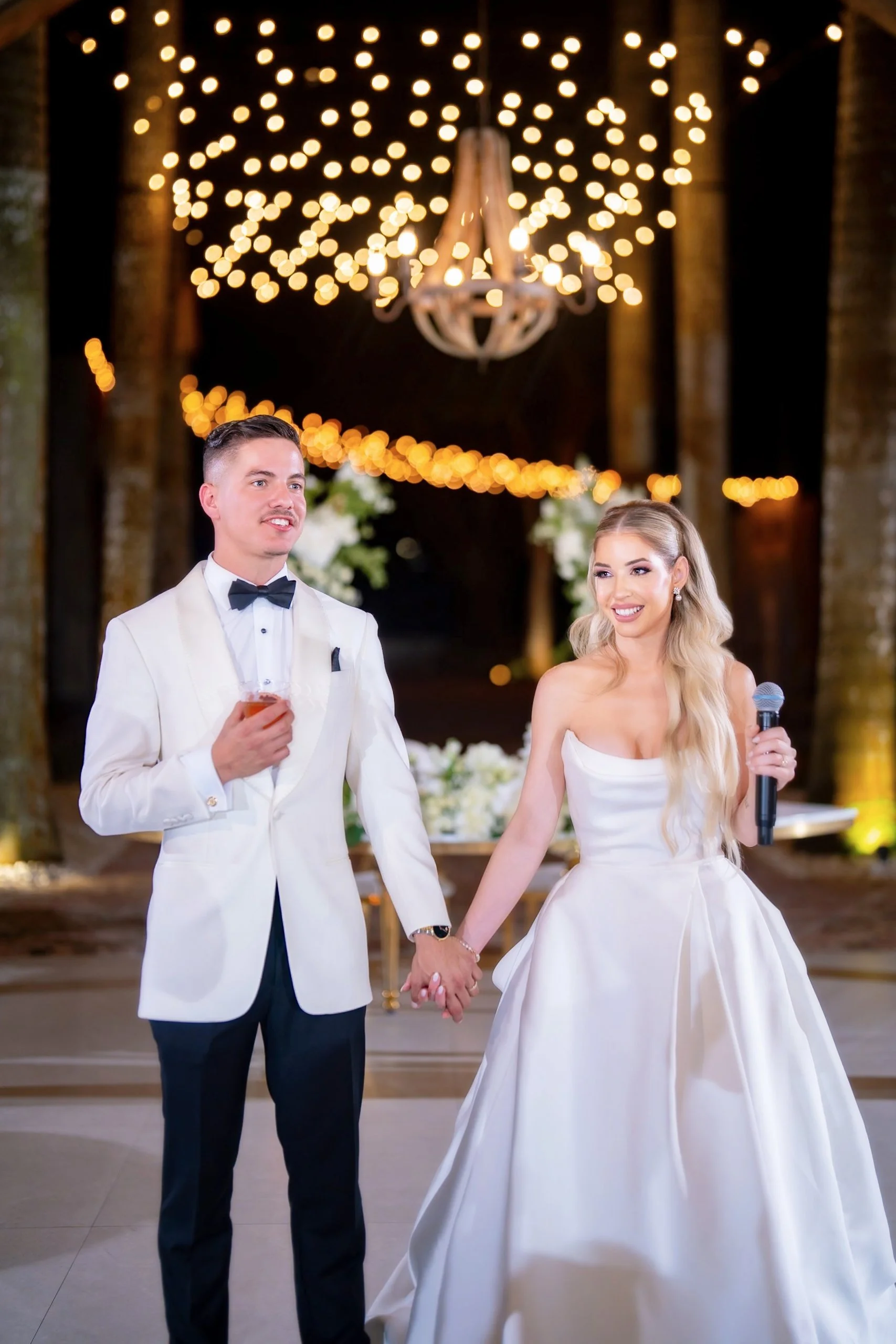 A bride and groom holding hands at their wedding reception, with the bride holding a microphone and the groom holding a drink, under warm string lights and a chandelier.