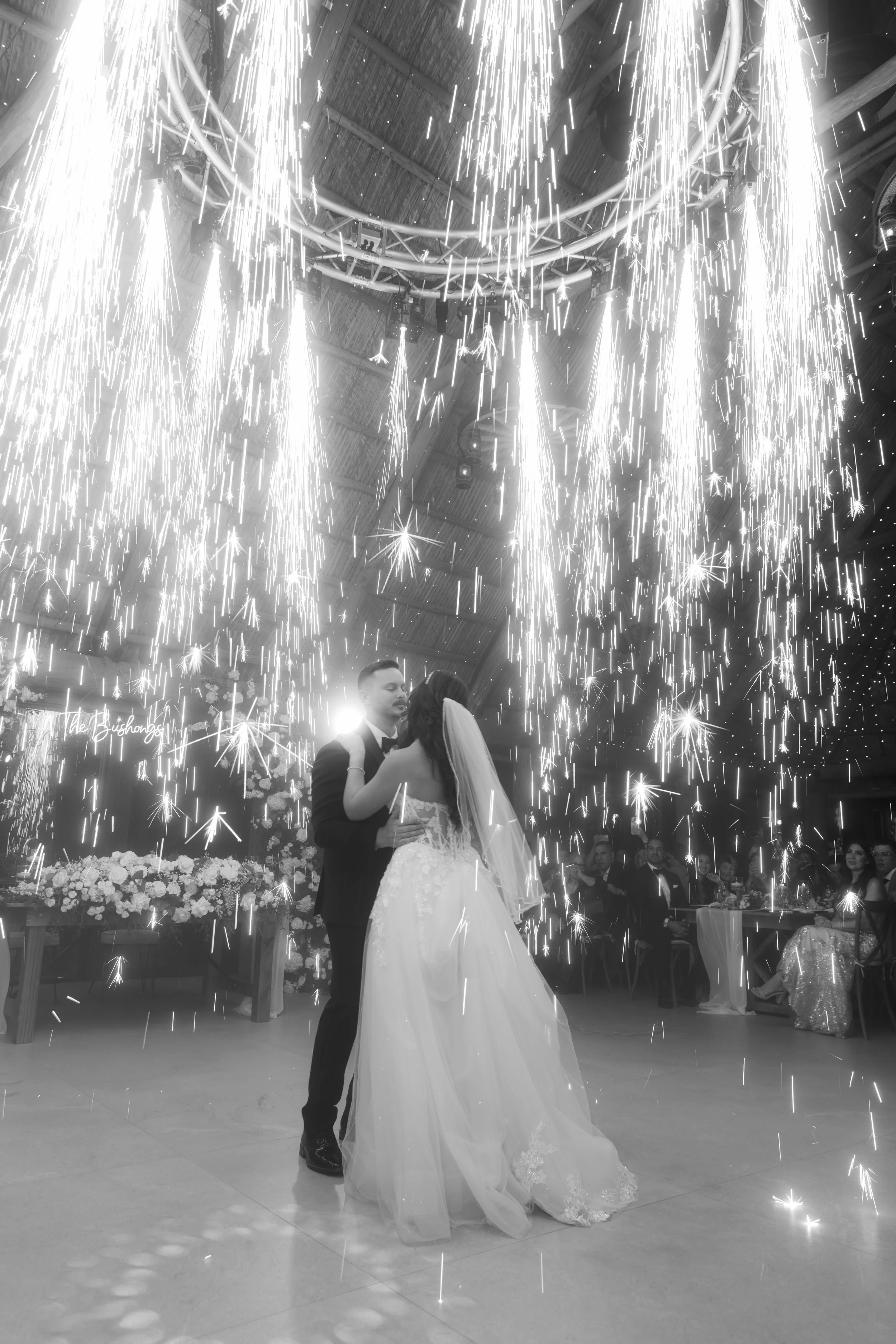 Bride and groom dancing under fireworks at their wedding reception, with guests watching in the background.