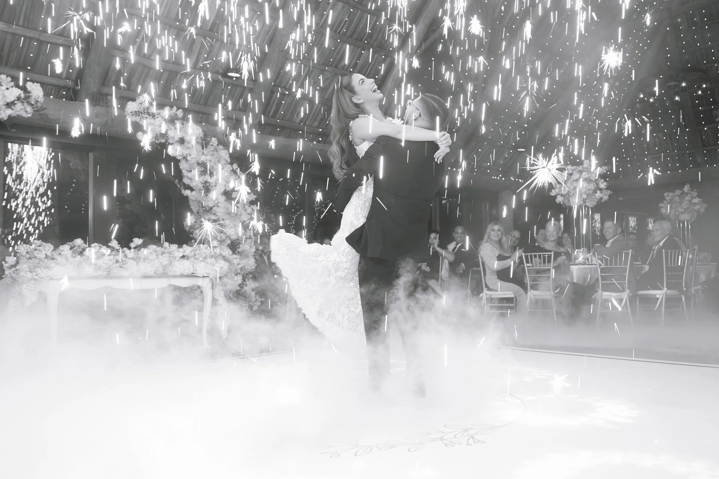 A couple dancing at their wedding reception, with sparks and smoke effects around them, guests seated at tables in the background.