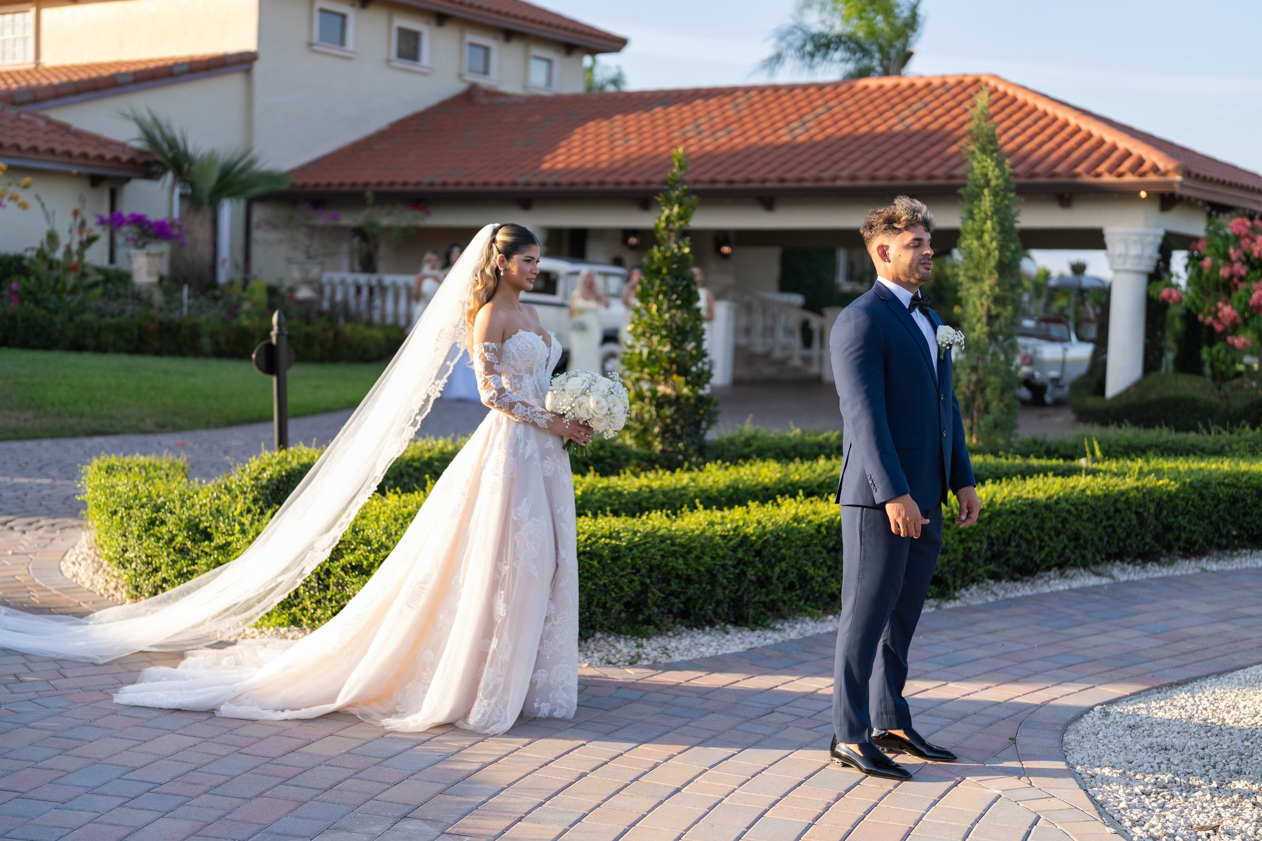 A bride in a wedding gown and a groom in a dark blue suit stand separately outdoors on a paved path during a wedding ceremony, with greenery and a house in the background.