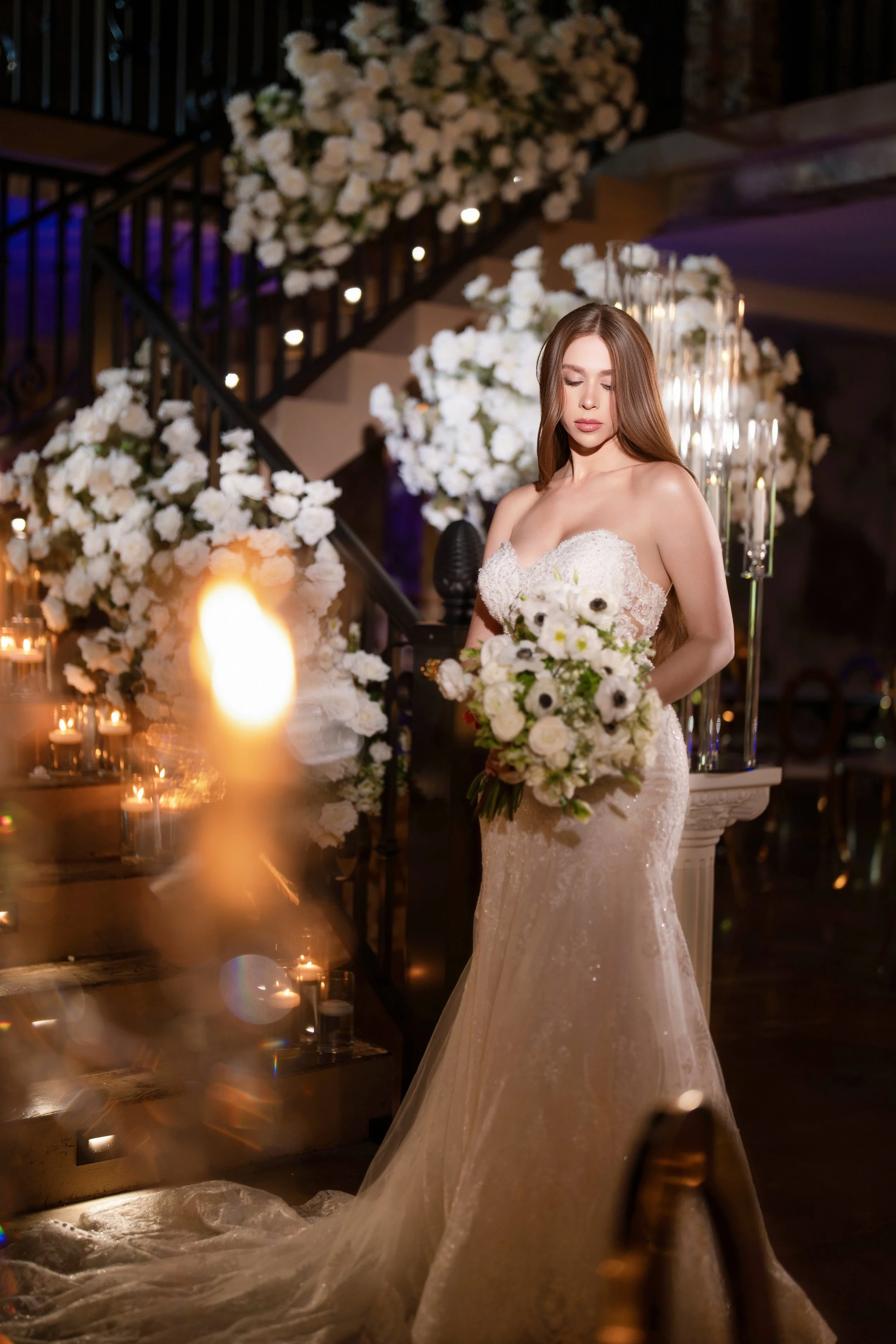 A bride in a strapless white wedding gown holding a bouquet of white flowers at a wedding reception with floral arrangements and candles in the background.