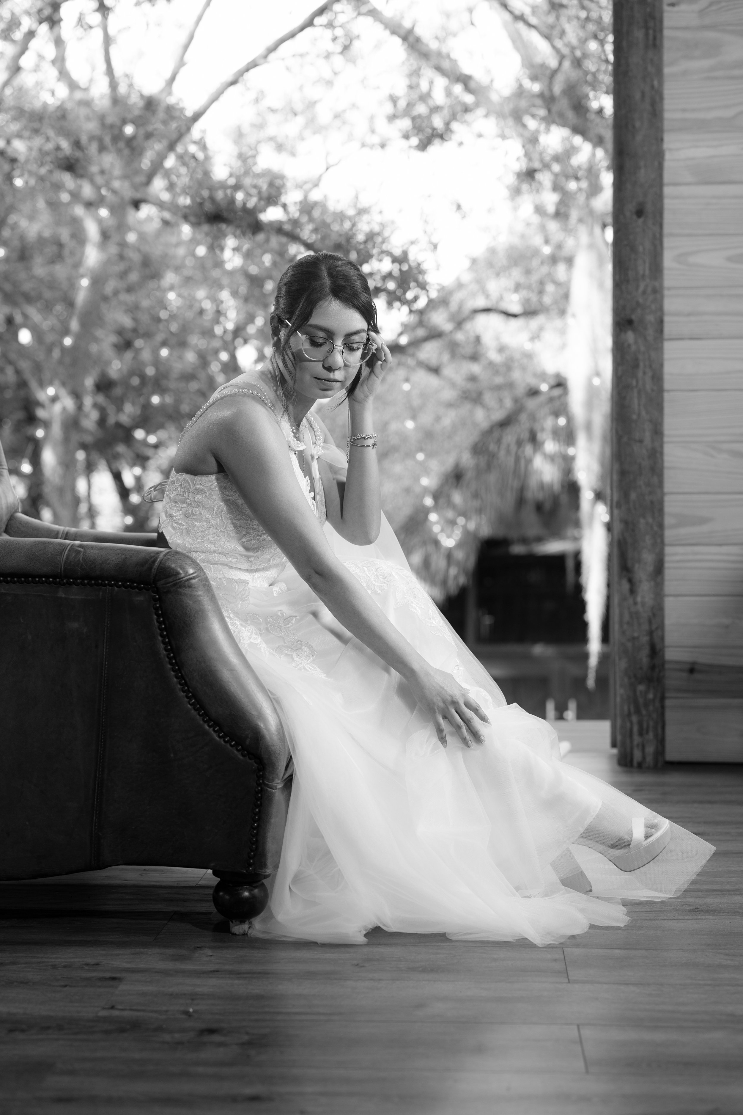A woman in a wedding dress and glasses sitting on a dark sofa, looking down and adjusting her dress, with trees visible outside through a window in the background.