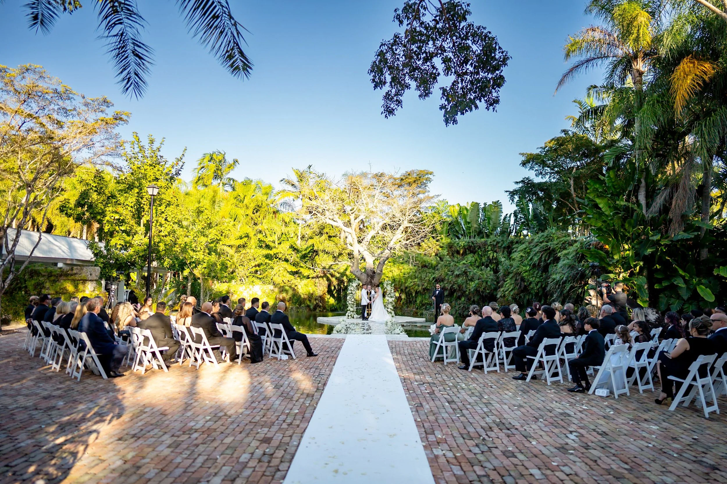 Outdoor wedding ceremony in a garden with white chairs arranged in rows on brick paving, a white aisle runner, a couple under a large tree with white flowers, surrounded by guests, lush greenery, and blue sky.