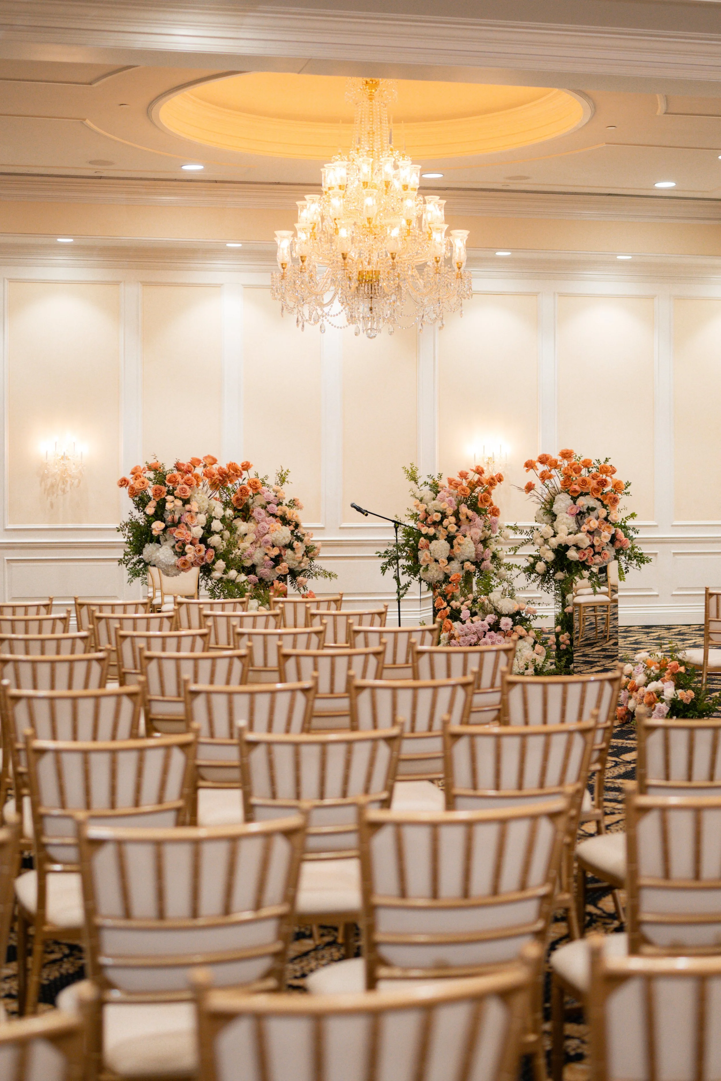 Elegant wedding ceremony setup with rows of gold chairs, floral arrangements, and a grand chandelier in a white ornate room.