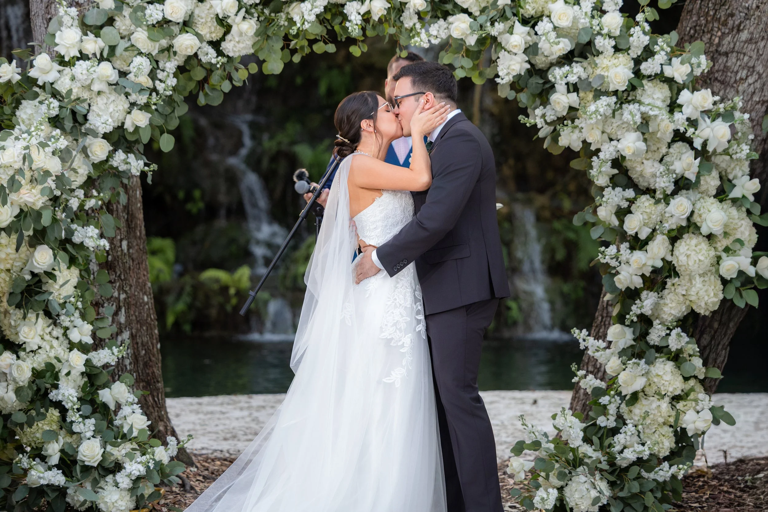 Bride and groom kissing during wedding ceremony, framed by a floral arch, with a waterfall in the background.