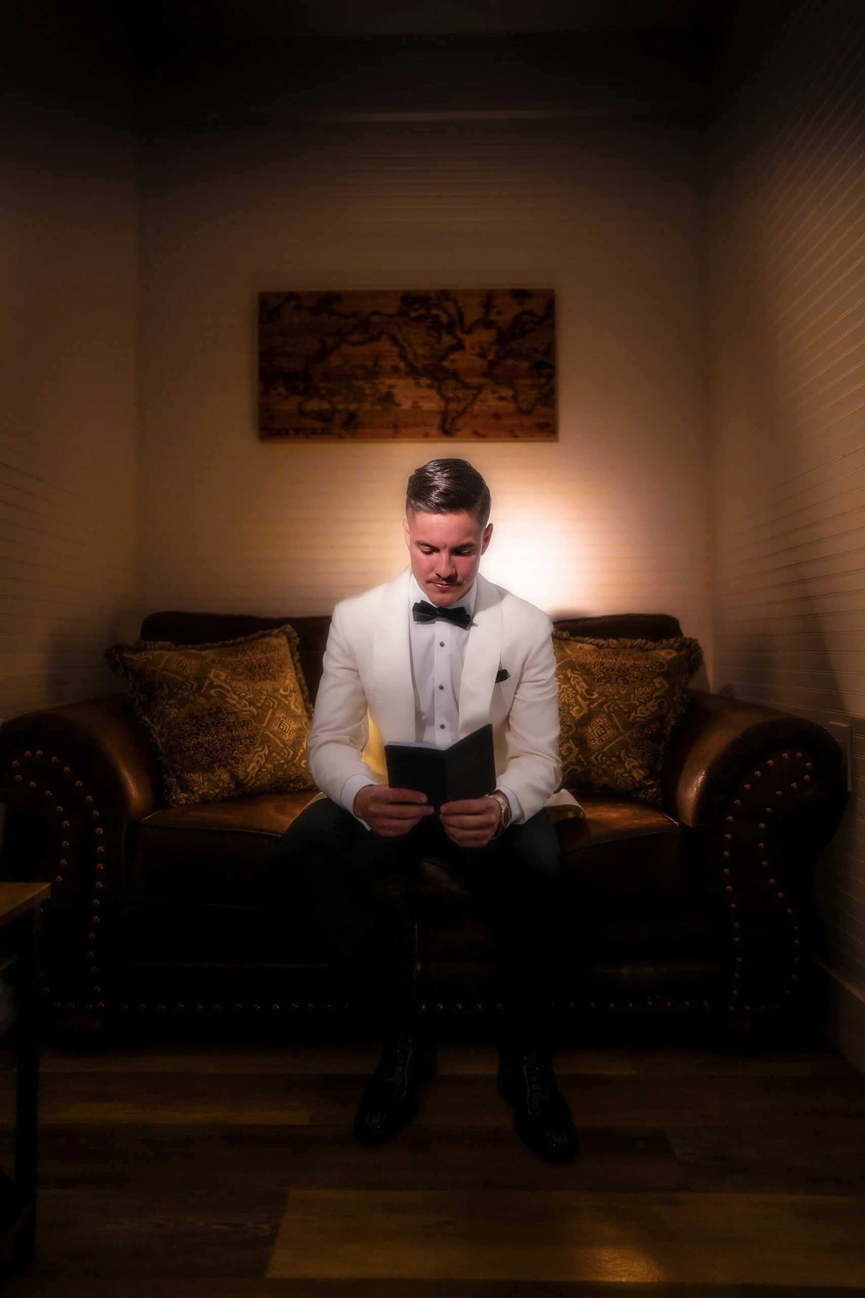 A man dressed in a white tuxedo jacket and black bow tie sitting on a dark leather sofa, reading a small book or booklet, in a warmly lit room with a world map hanging on the wall behind him and decorative pillows.