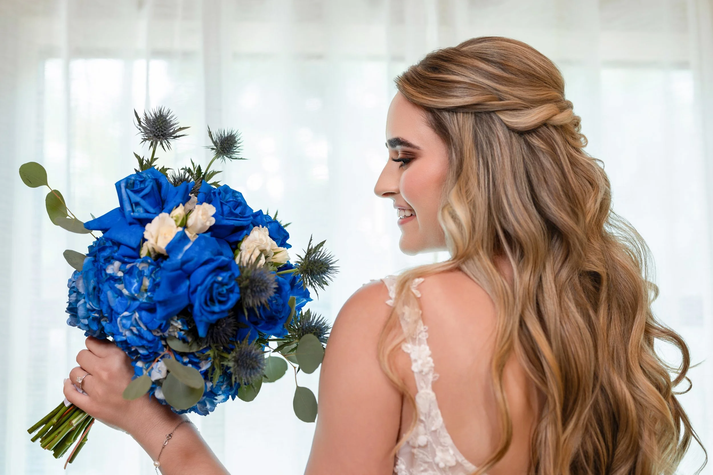 A woman with long wavy hair holding a bouquet of blue and white flowers, smiling, with a blurred background of a light-colored curtain or window.