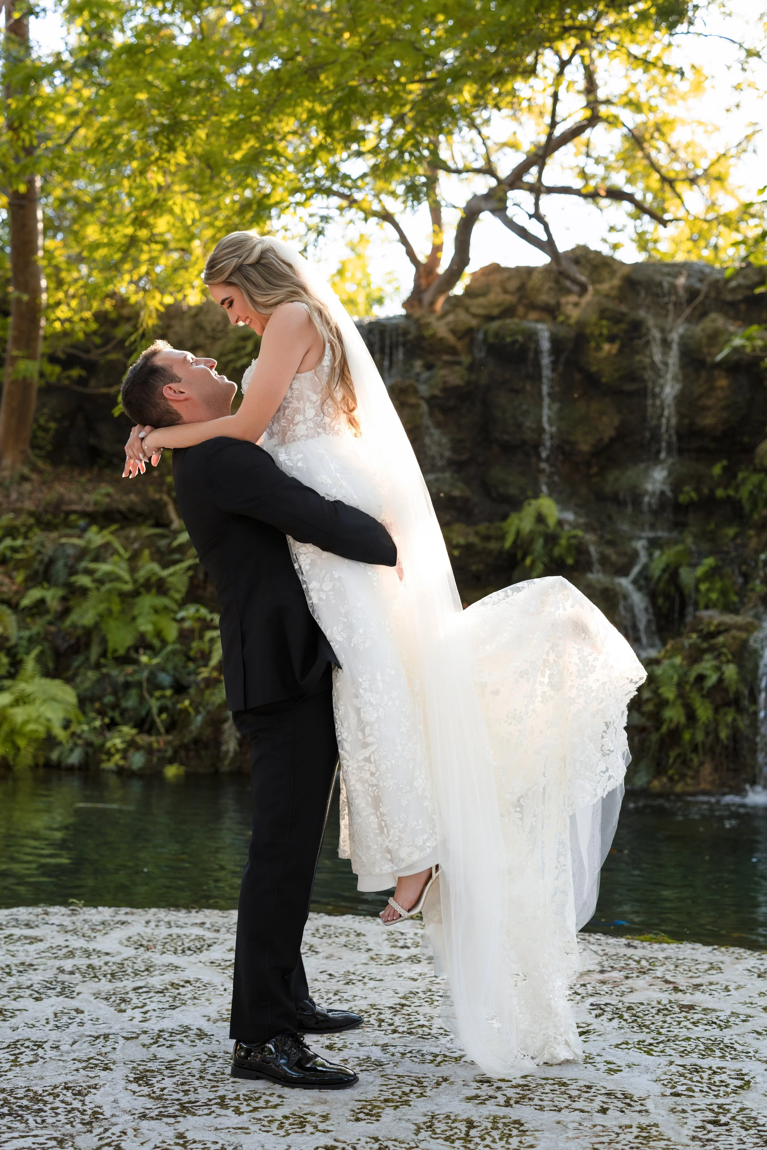 A bride and groom in a joyful embrace outdoors near a waterfall, with the groom lifting the bride in a wedding dress.