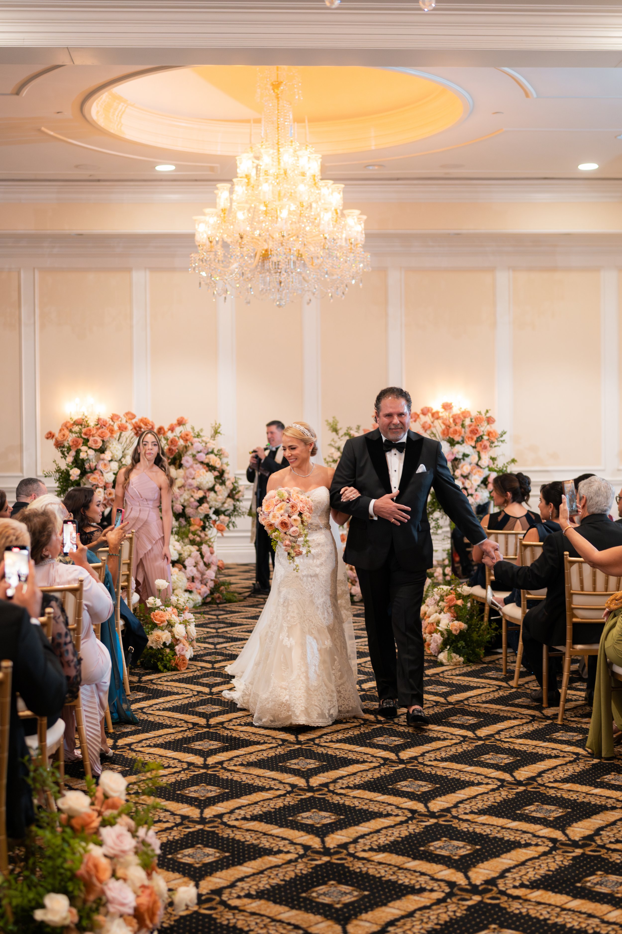 A bride walking down the aisle with her father at a wedding reception. The ballroom has a chandelier, floral arrangements, and seated guests taking photos.