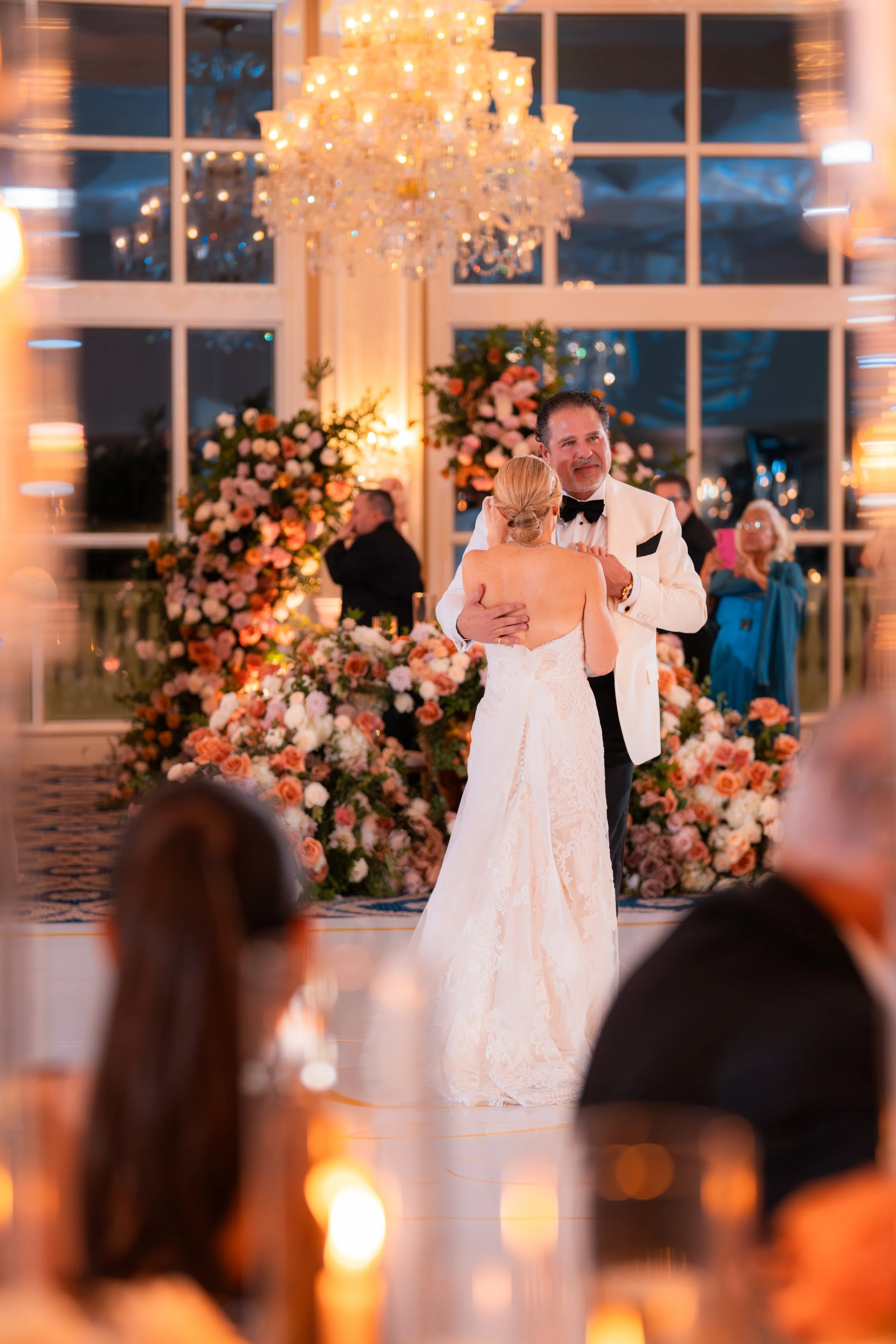 A bride and groom dancing at their wedding reception, with guests observing in the background, elegant floral arrangements, and a large chandelier overhead in a bright, decorated venue.