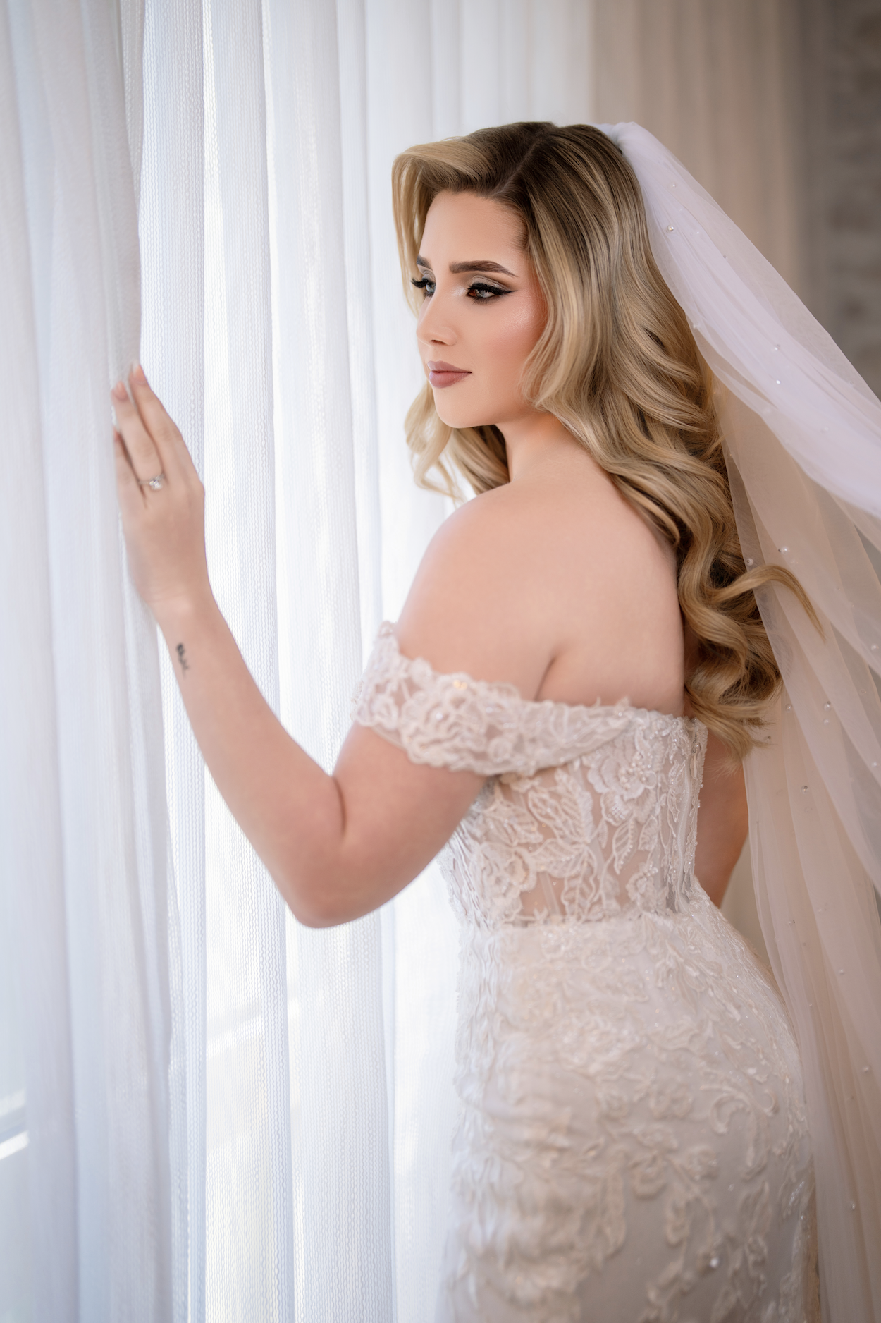 A bride with wavy blonde hair in a strapless lace wedding gown, standing by a sheer curtain and looking outside.