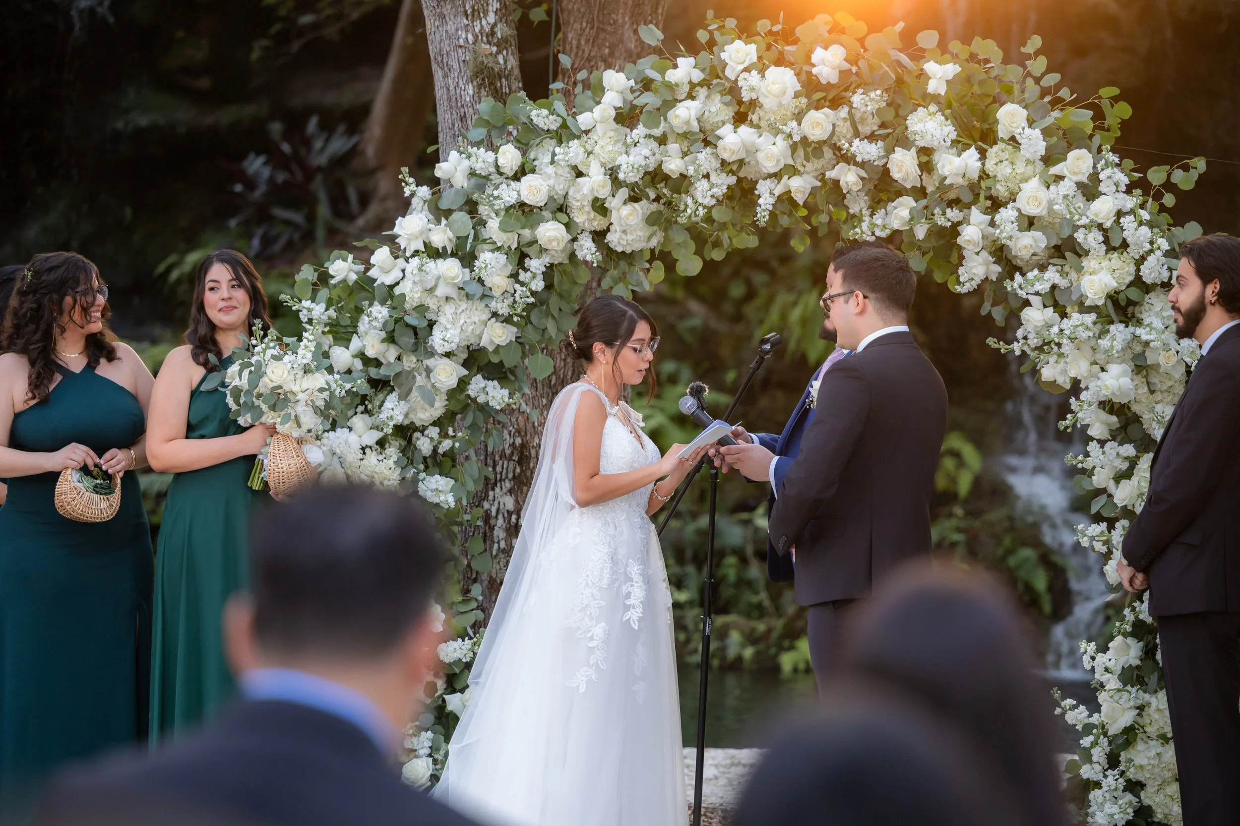 A wedding ceremony taking place outdoors near a body of water. The bride and groom are facing each other, holding hands, with the officiant reading from a book. The bride is in a white lace wedding dress with a veil, and the groom is in a dark suit. 