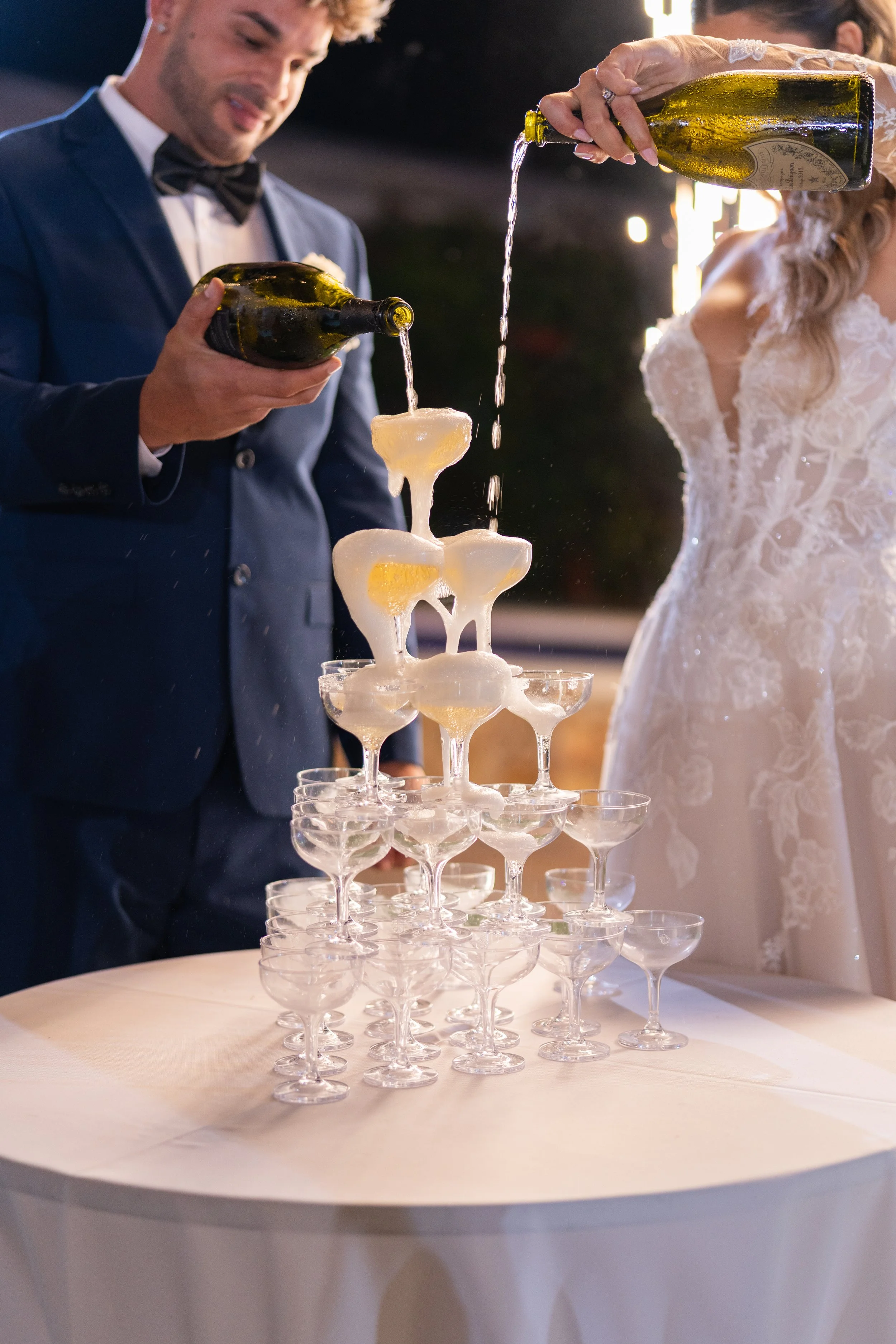 Couple pouring champagne into a pyramid of glasses with foam overflowing, at a celebration event.
