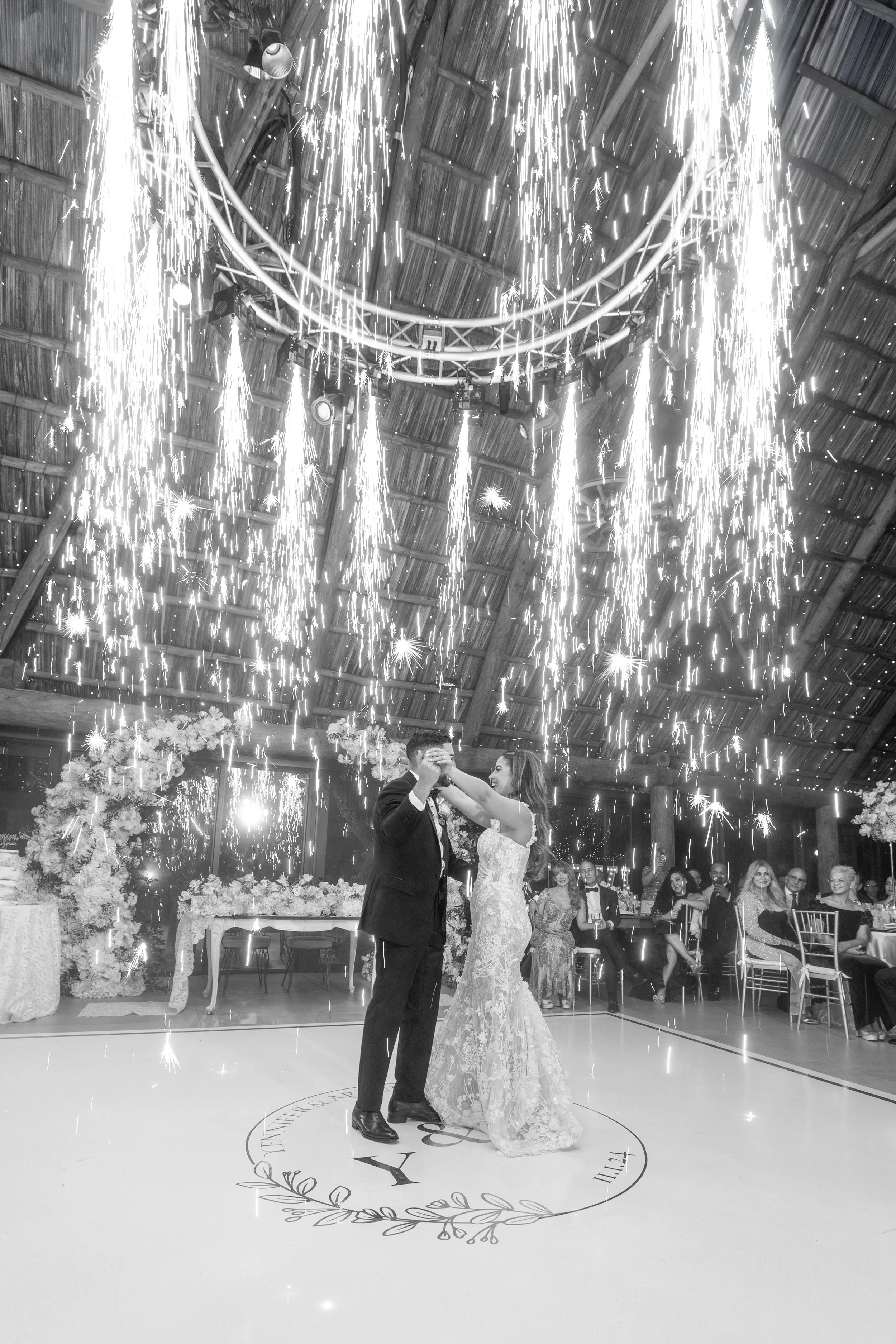 A bride and groom dancing during their wedding reception under a fireworks display, with guests seated around them in a rustic wooden venue decorated with flowers.