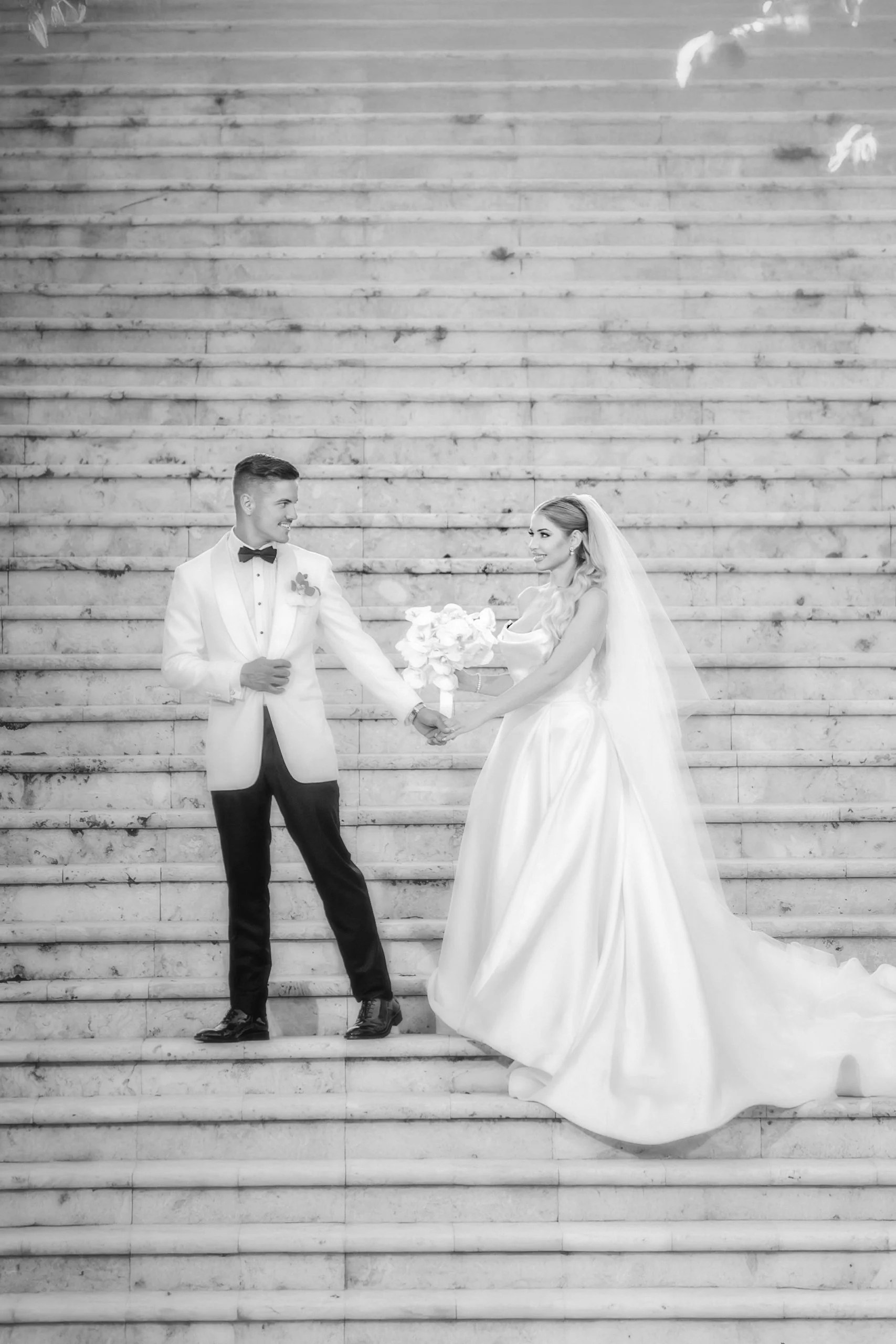 A bride and groom stand on steps, holding hands and facing each other, with the bride holding a bouquet. The groom is dressed in a tuxedo with a bow tie, and the bride is in a long wedding gown with a veil.