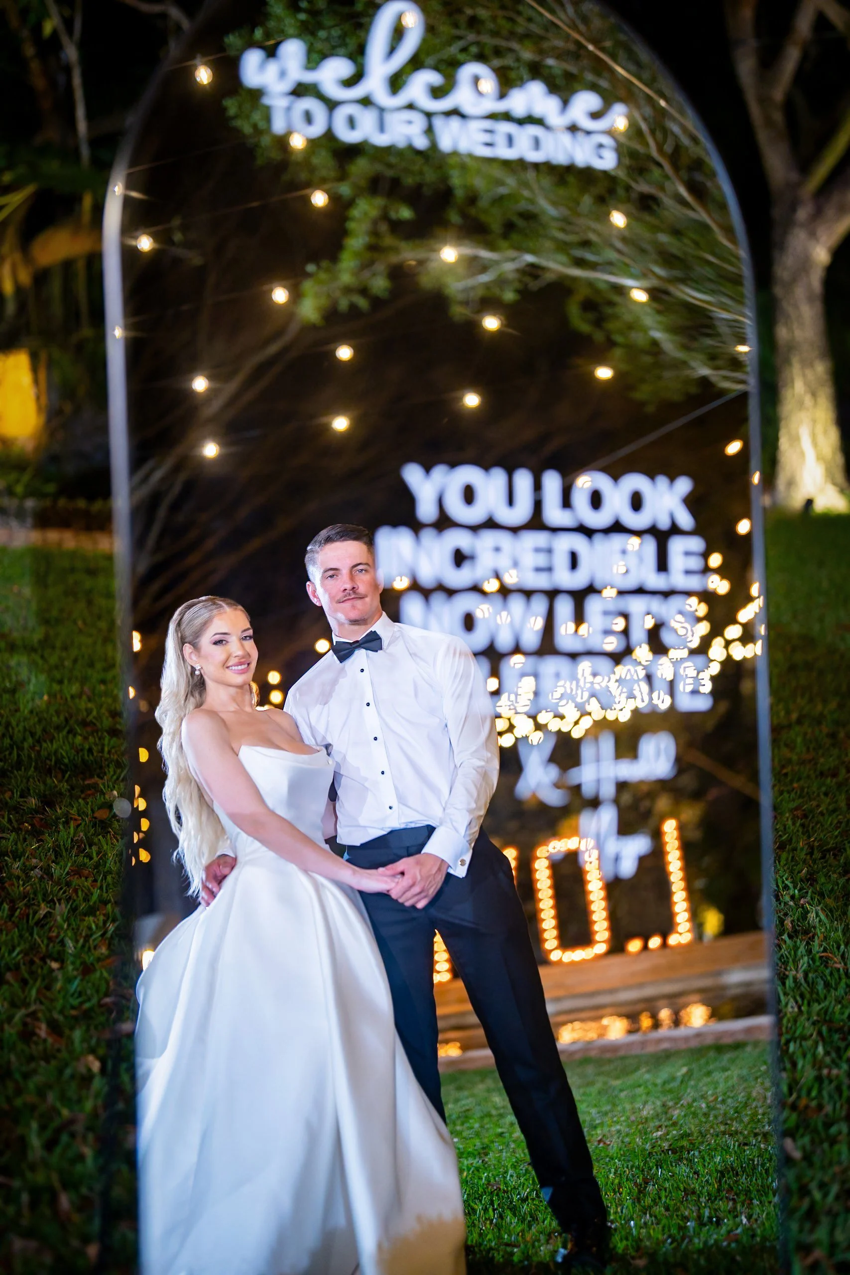 A newlywed couple posing outdoors at night, reflected in a large mirror with glowing wedding messages and lights, with greenery and a tree in the background.