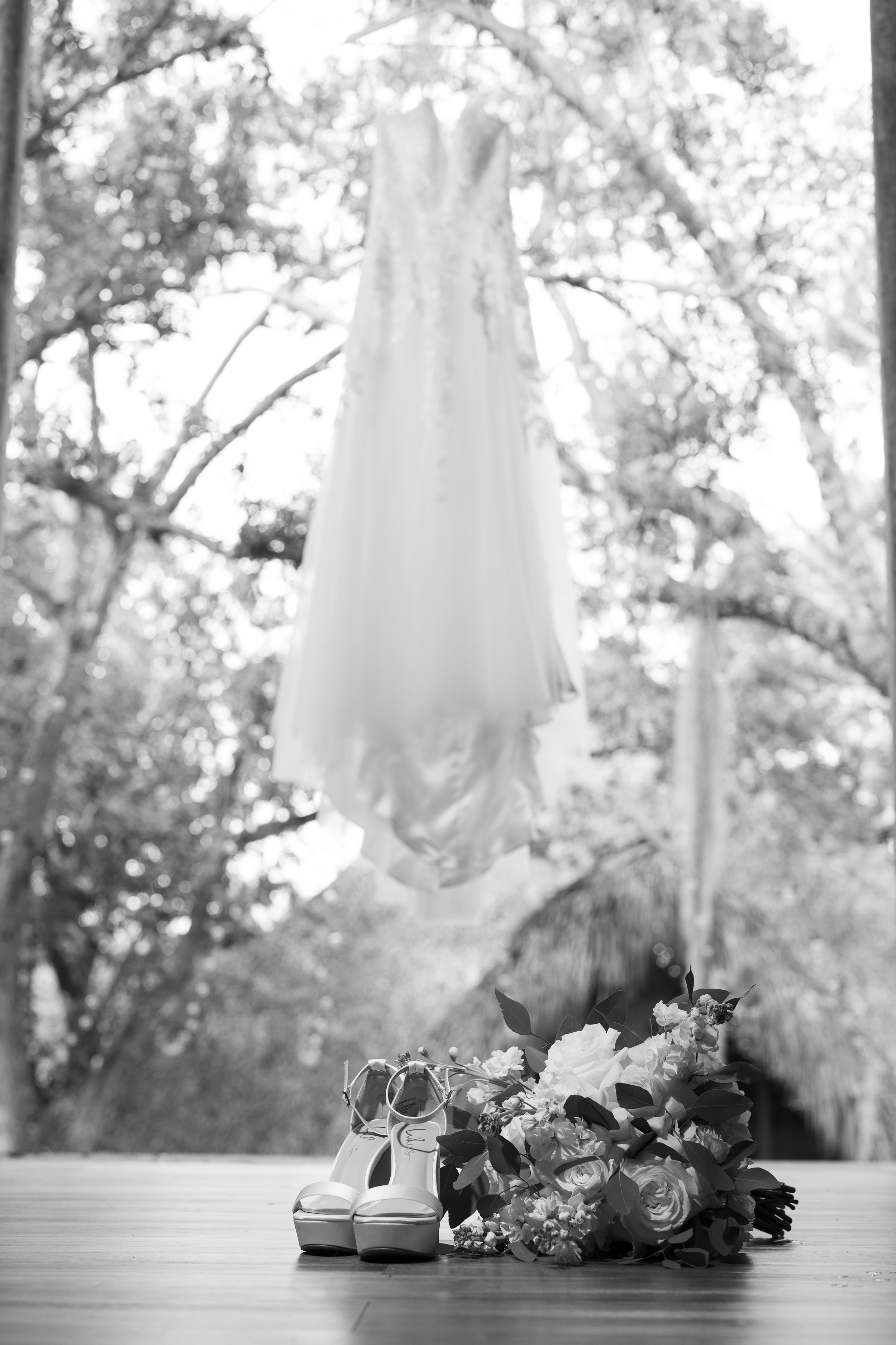 Bride's shoes and bouquet on a wooden floor with a dress hanging from a tree and tropical trees in the background.
