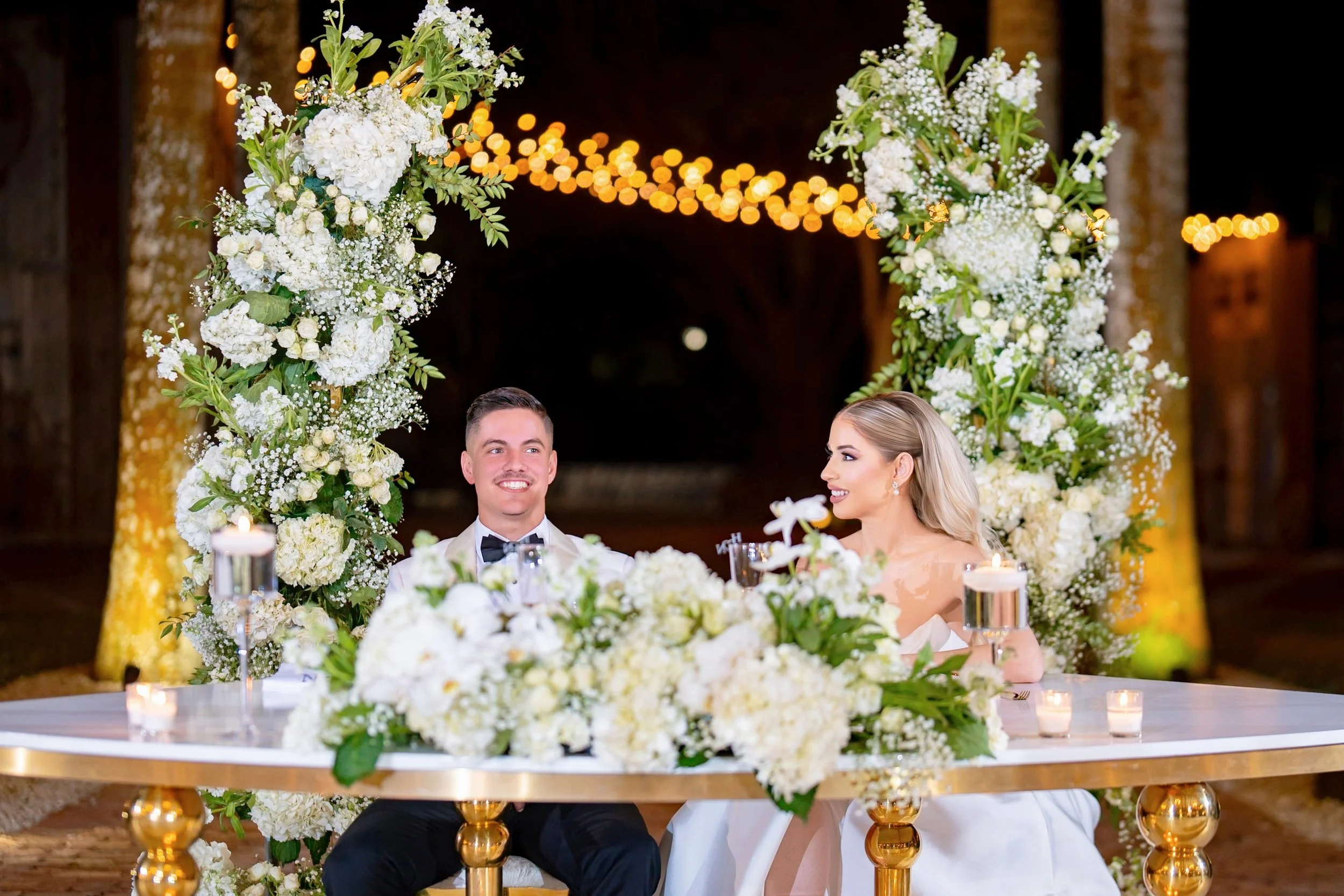 Bride and groom sitting at a decorated table with white flowers, candles, and greenery, under a floral arch with hanging lights during a wedding reception at night.