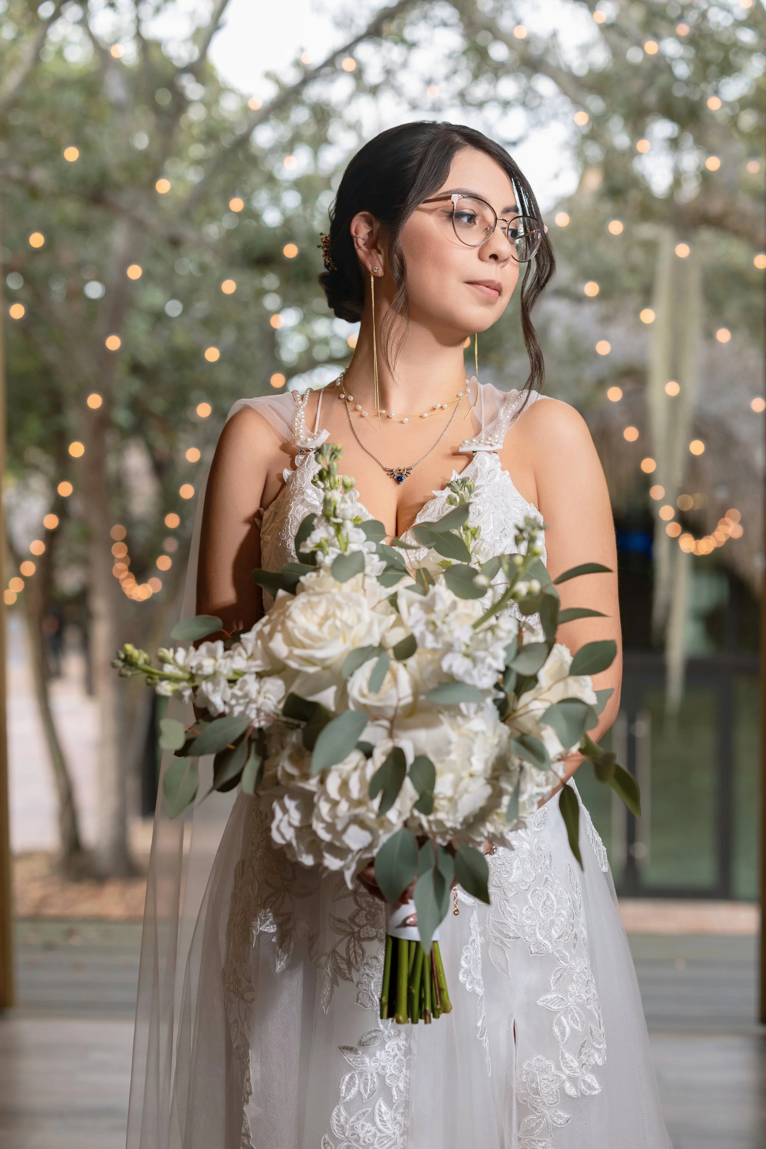 A young woman in a white wedding dress holds a bouquet of white flowers and greenery, standing outdoors with string lights and trees in the background.