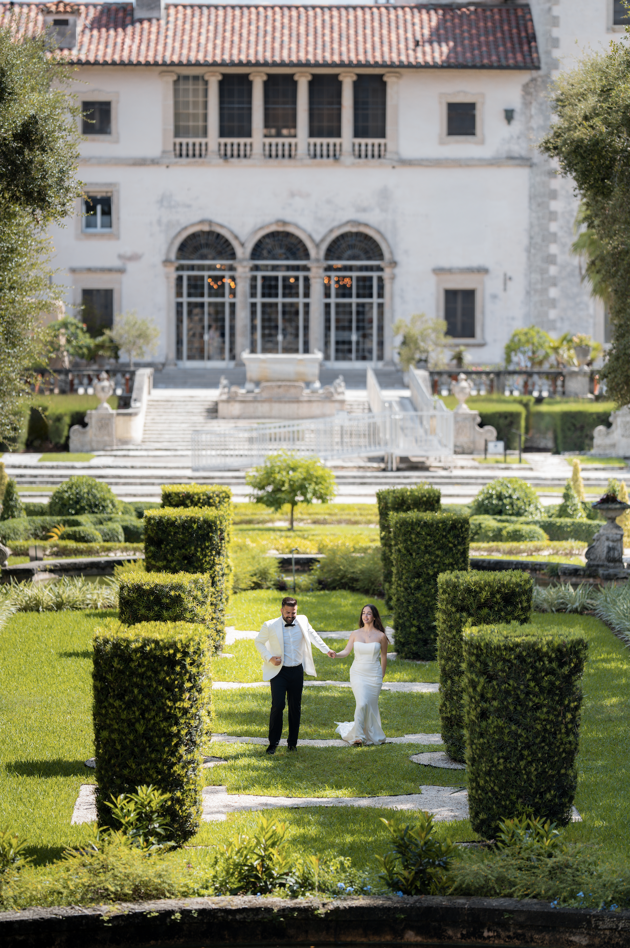 A bride and groom holding hands in a formal garden with a historic mansion in the background.