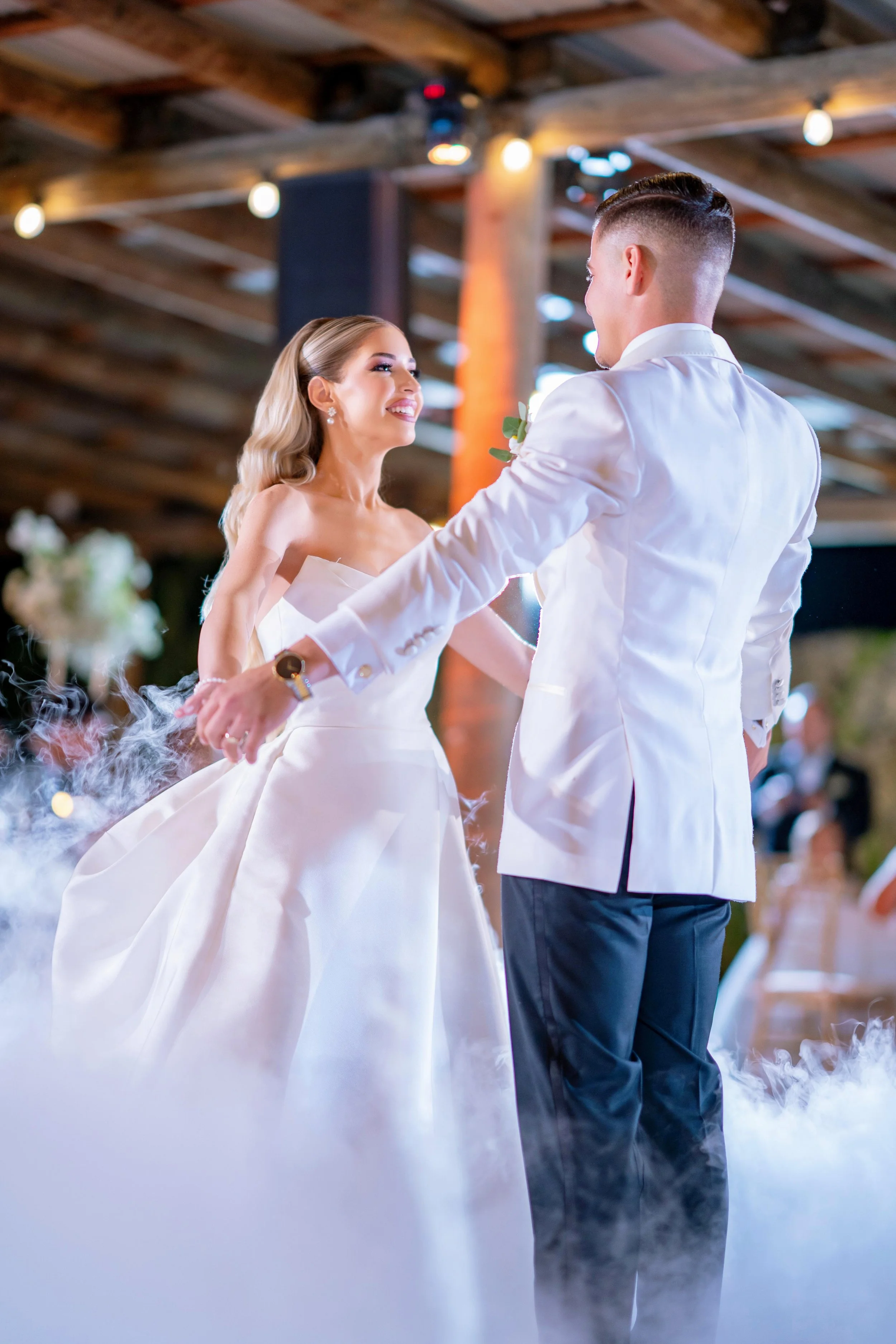 A bride and groom dancing at their wedding reception, with smiling faces, in a rustic indoor venue with warm lighting and wooden beams, surrounded by fog and blurred guests.