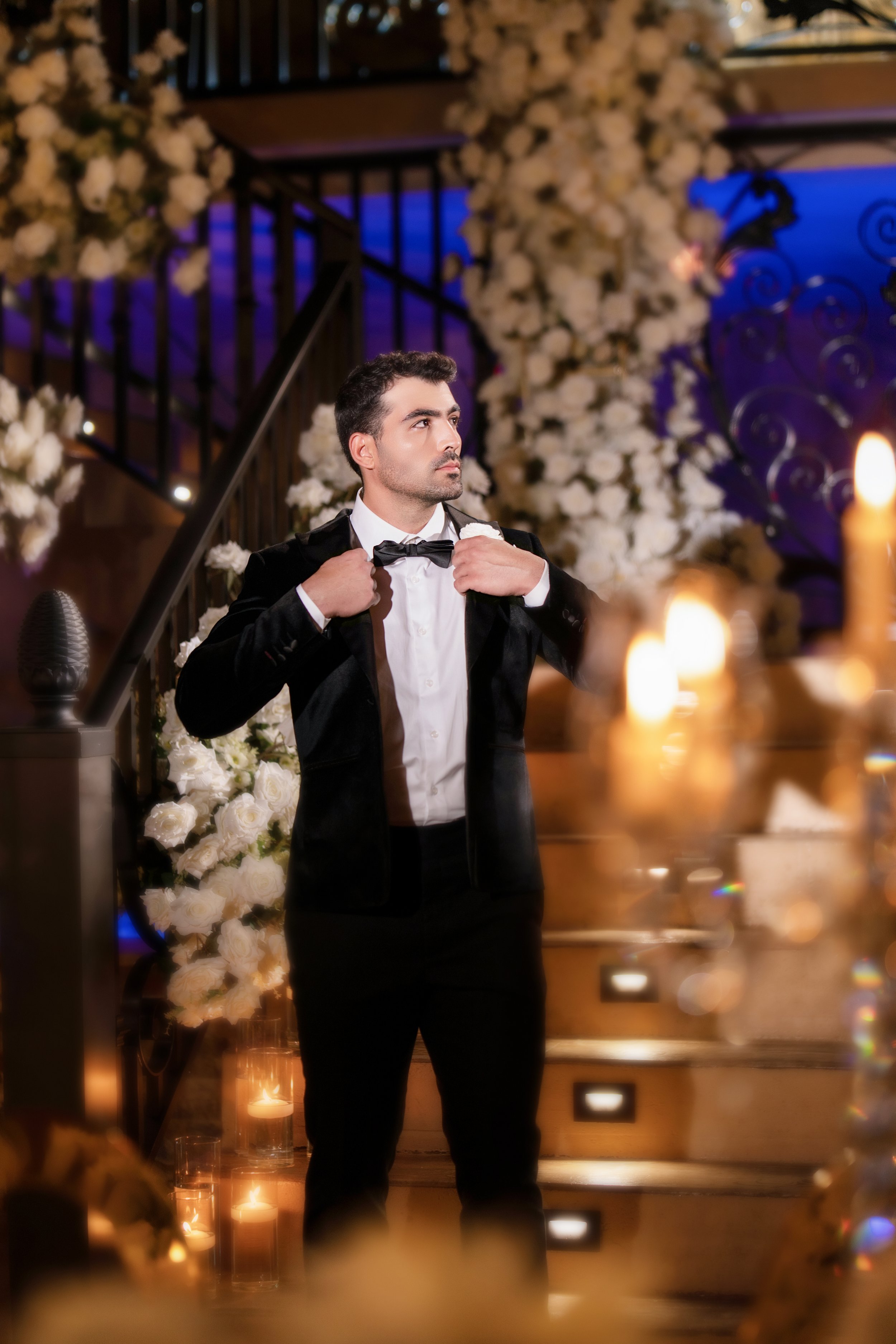 A man in a tuxedo adjusting his bow tie at a formal event with floral decorations and candles.