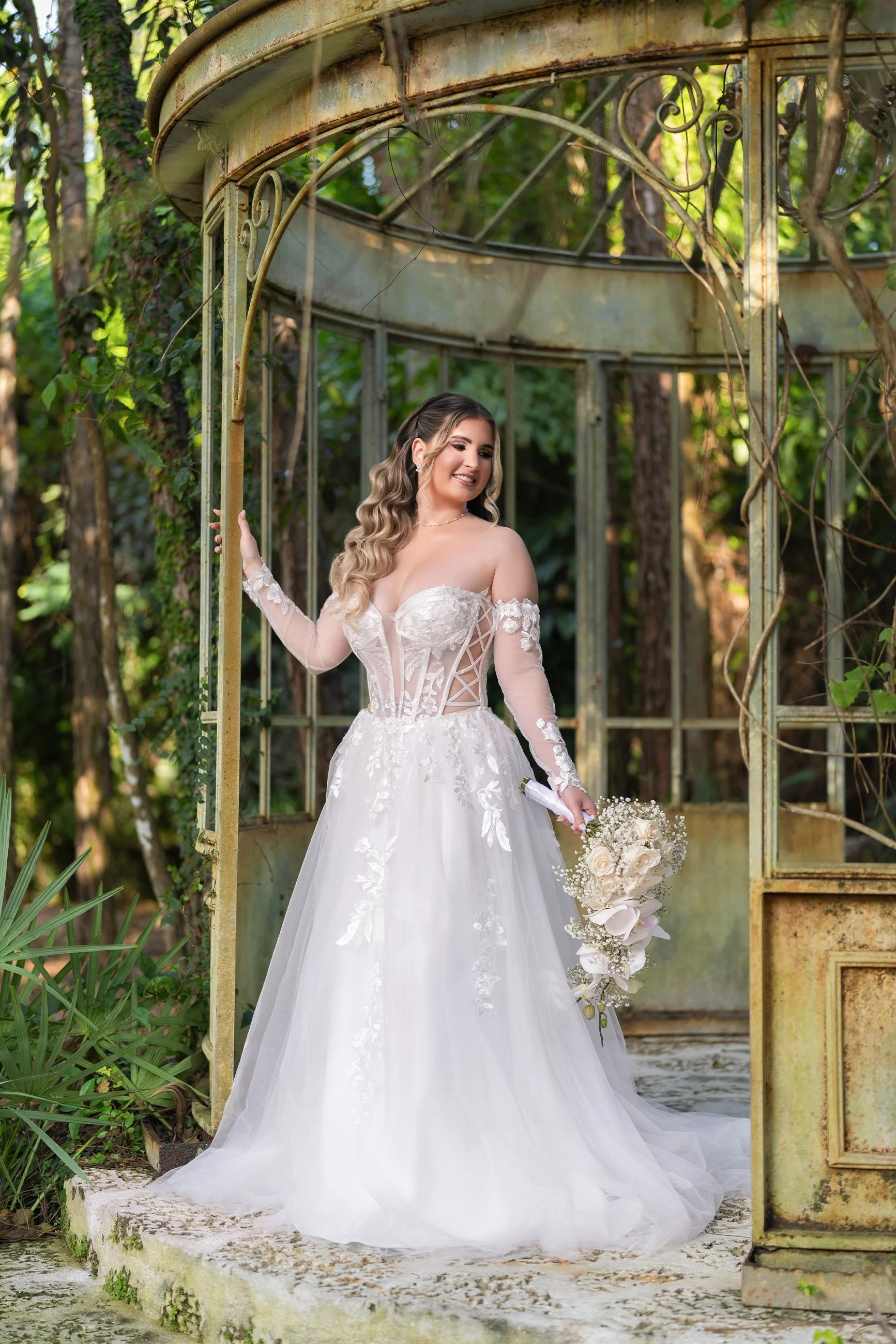 A bride in a white wedding gown with lace details, standing under a rustic, weathered metal gazebo in a lush, green outdoor setting, holding a bouquet of white roses and greenery.