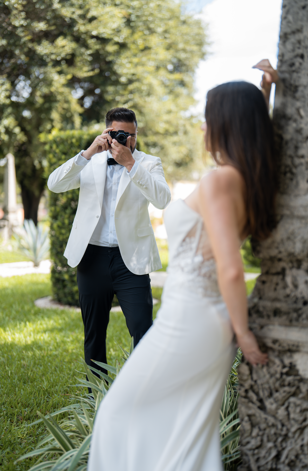A man in a white tuxedo jacket taking a photo of a woman in a wedding dress leaning against a tree in a park.