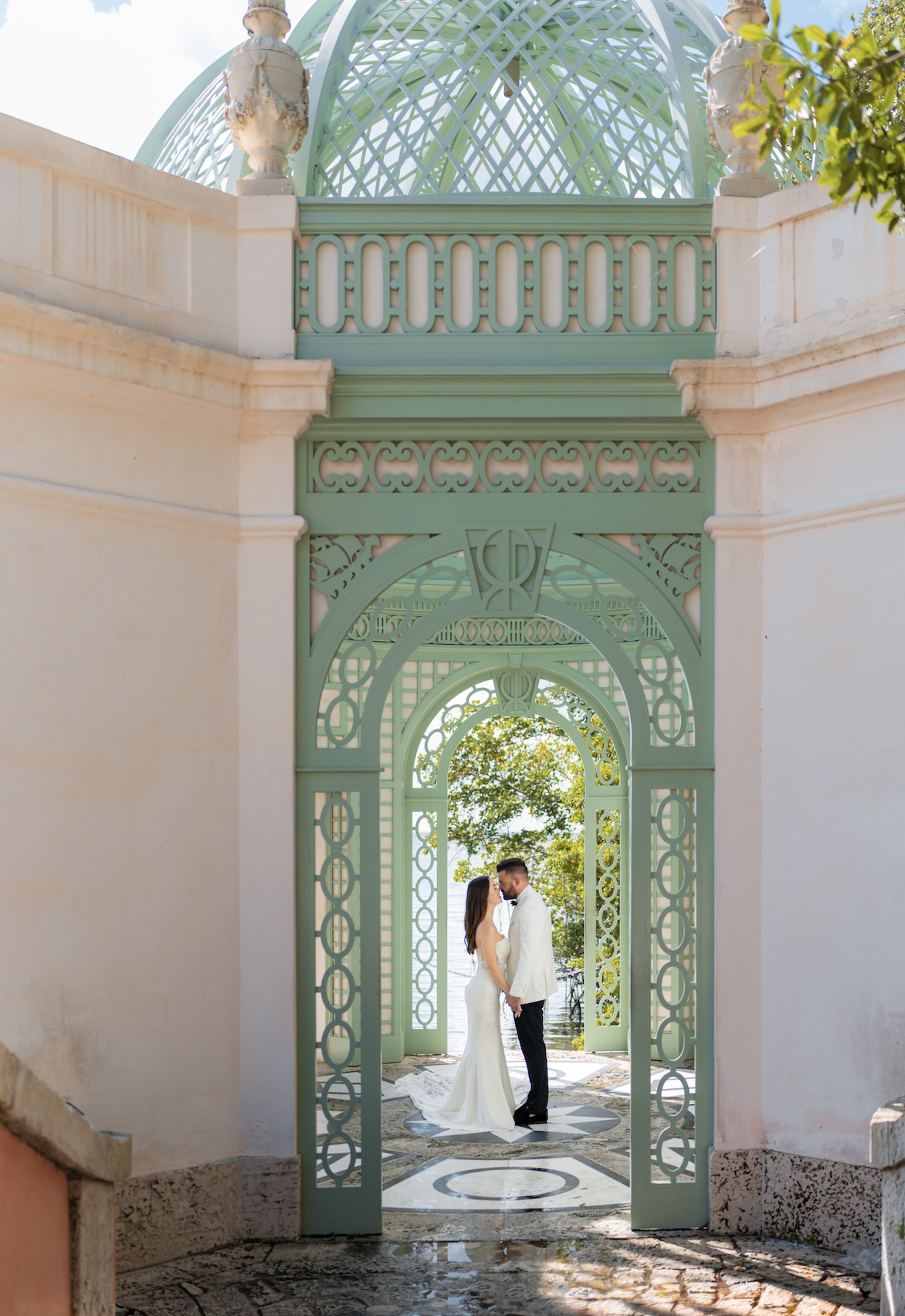 A couple in wedding attire standing face to face and holding hands in a decorative arched gateway with ornate green ironwork, overlooking a tree and the water in the background.