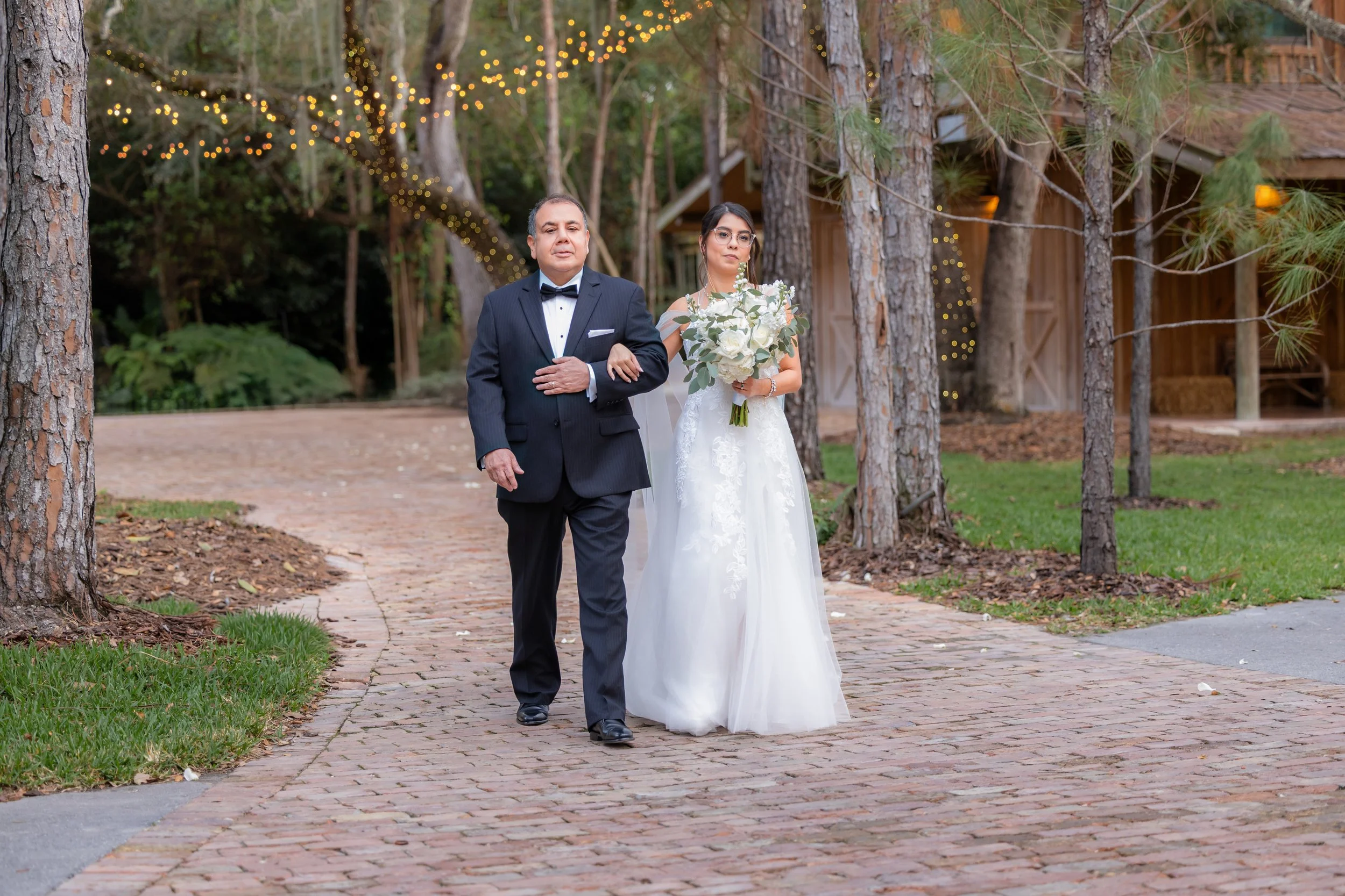 A bride walking down a garden pathway, arm in arm with a man in a tuxedo, holding a bouquet of white flowers under string lights and surrounded by trees.