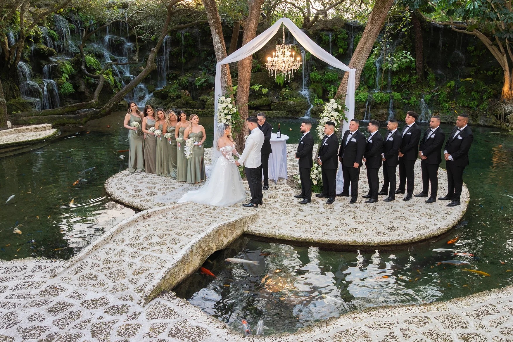 Wedding ceremony with bride and groom holding hands, standing on a stone platform surrounded by water, with bridesmaids and groomsmen in formal attire, set outdoors in a lush green garden with waterfalls and trees.
