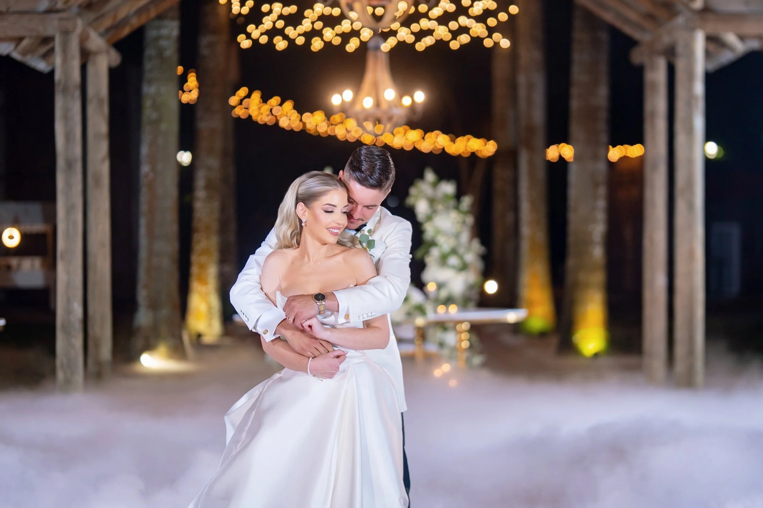 A bride and groom embracing on a dance floor at their wedding reception, with warm string lights and a floral arrangement in the background.