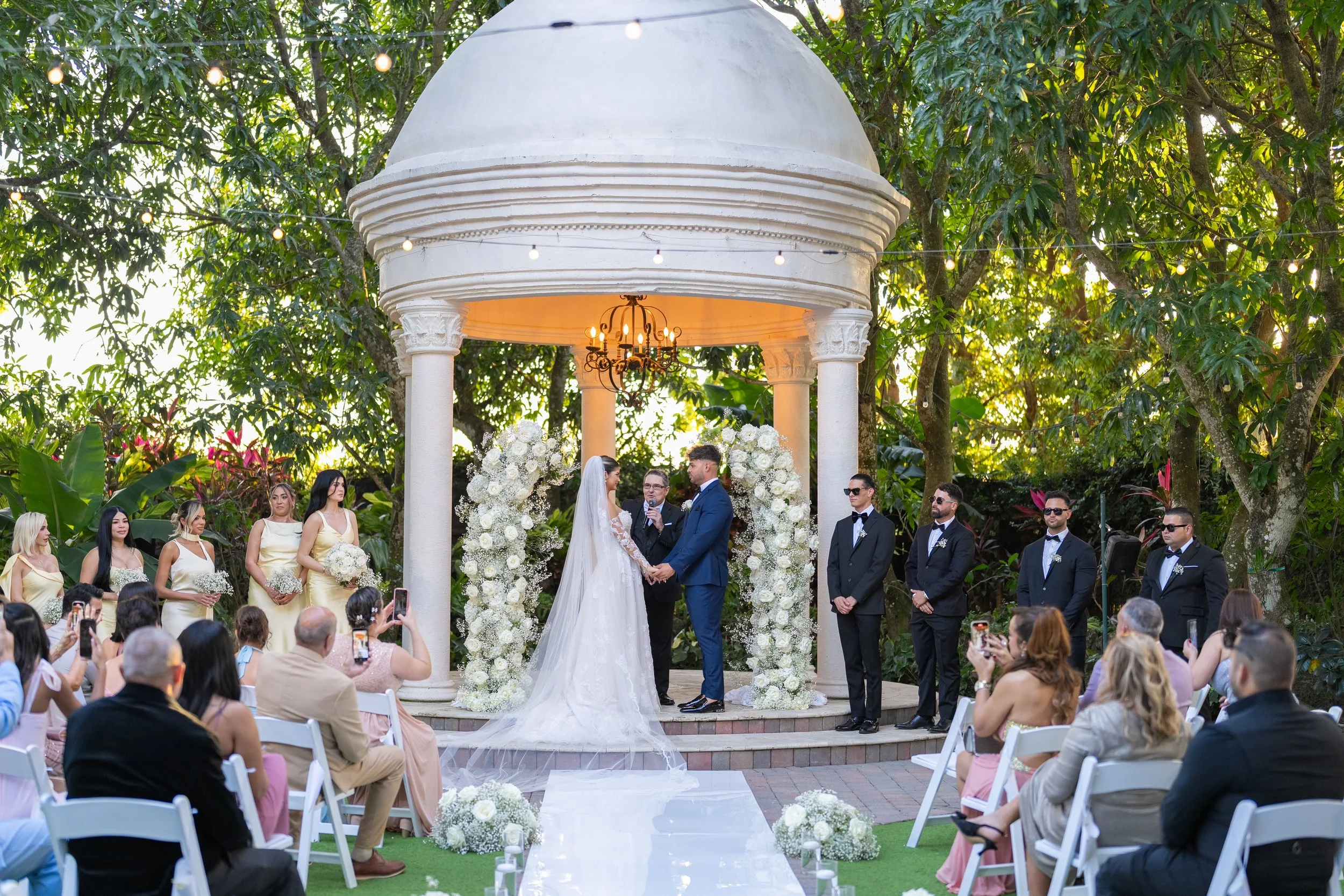 A wedding ceremony outdoors under a small white pavilion with columns, surrounded by trees. The bride and groom are holding hands and facing each other, with the officiant between them. The bride is in a white wedding gown with a long veil, and the g