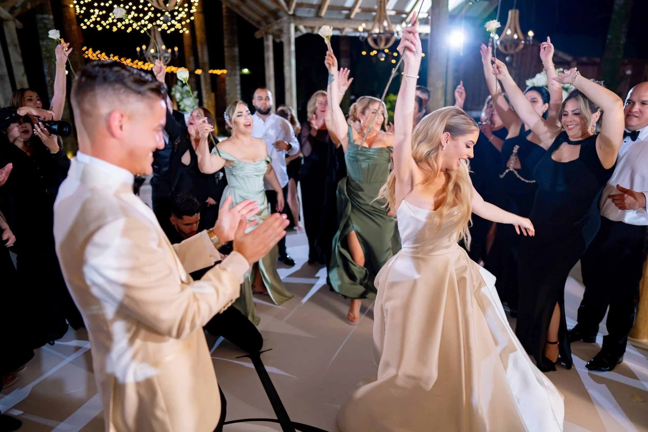 People dancing and celebrating at a wedding reception, with the bride in a white dress and the groom clapping, surrounded by guests raising their hands and smiling under string lights.