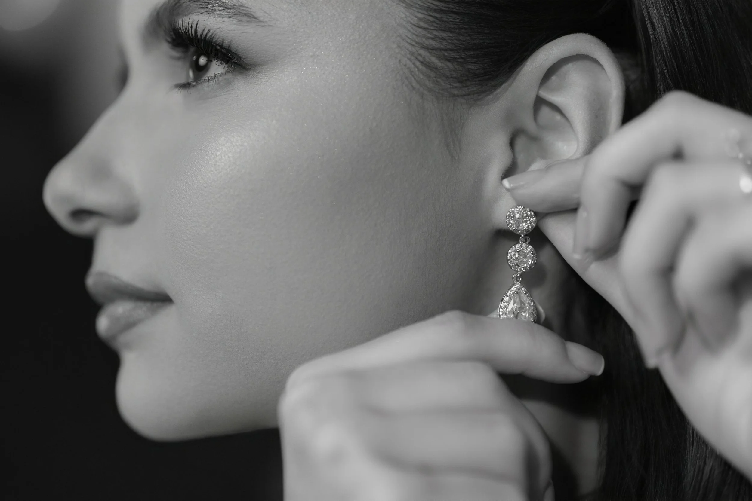 A black and white close-up of a woman adjusting an ornate dangling earring adorned with multiple diamonds.