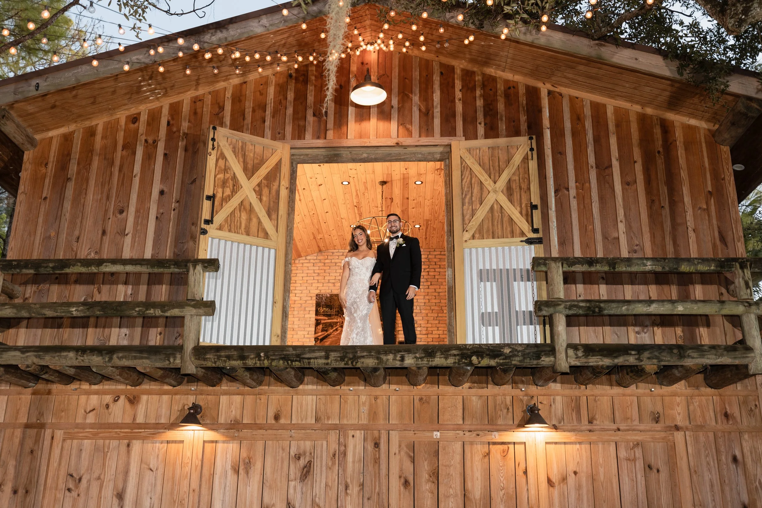 A bride and groom standing inside an open barn door on a wooden balcony with string lights hanging above, brick interior walls, and wooden ceiling, celebrating a wedding.