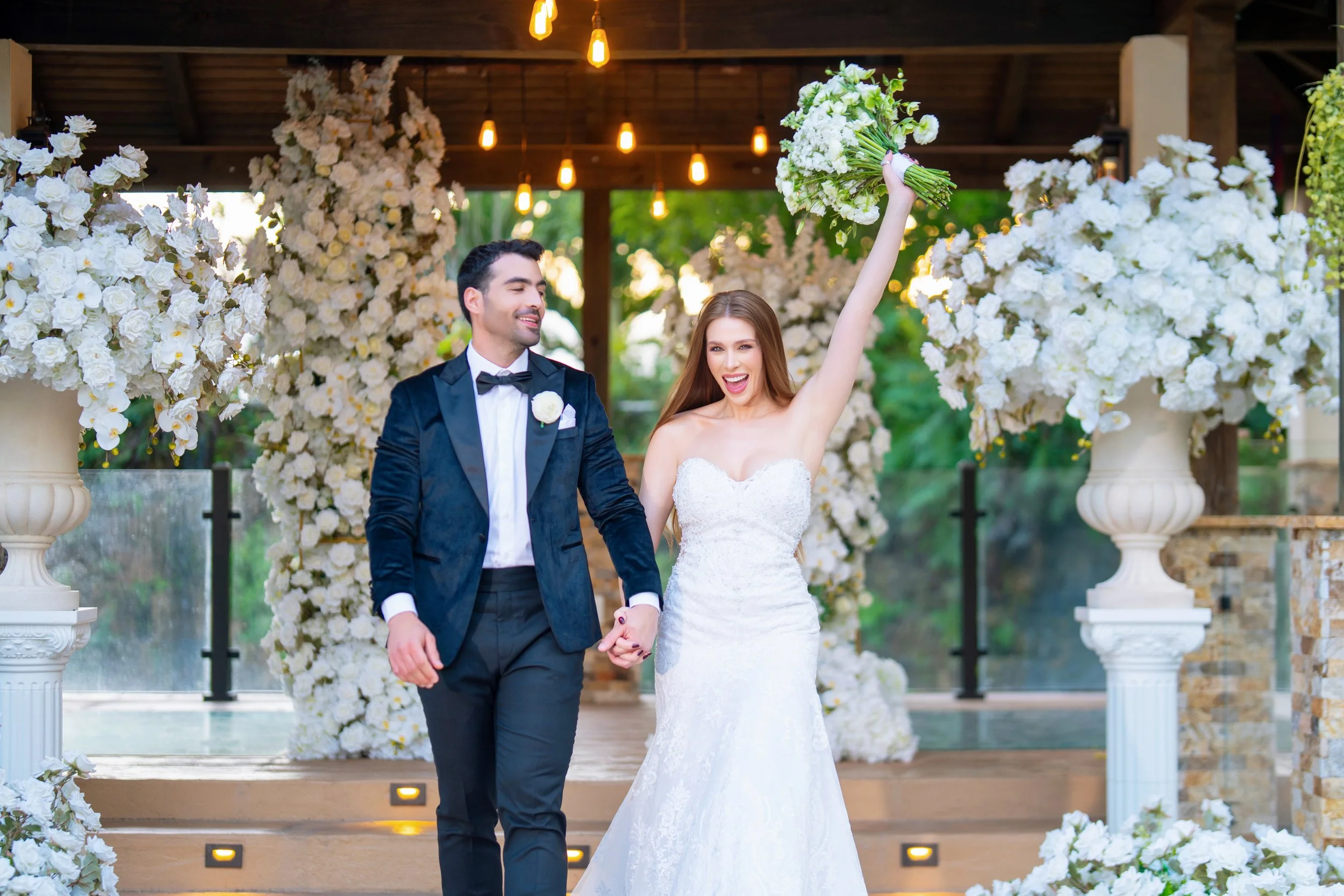 The bride and groom celebrating at their wedding, with the bride holding a bouquet in the air, standing in front of a floral arch decorated with white flowers.