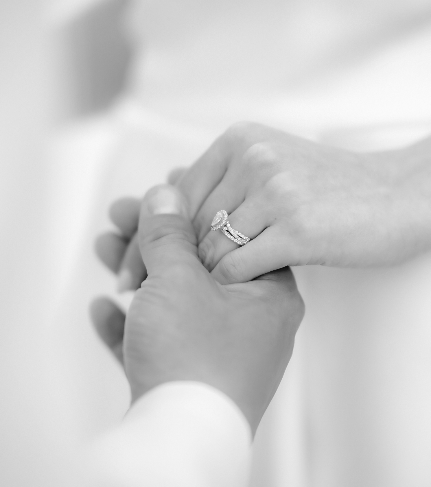 Close-up of a woman's hands, wearing an engagement ring and wedding band, holding a man's hand, in black and white.
