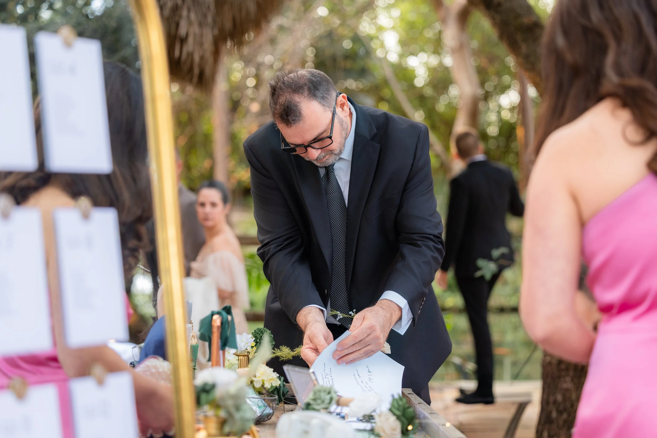 A man in a black suit with glasses appears to be signing a document at a wedding or outdoor event surrounded by women and decorated tables.