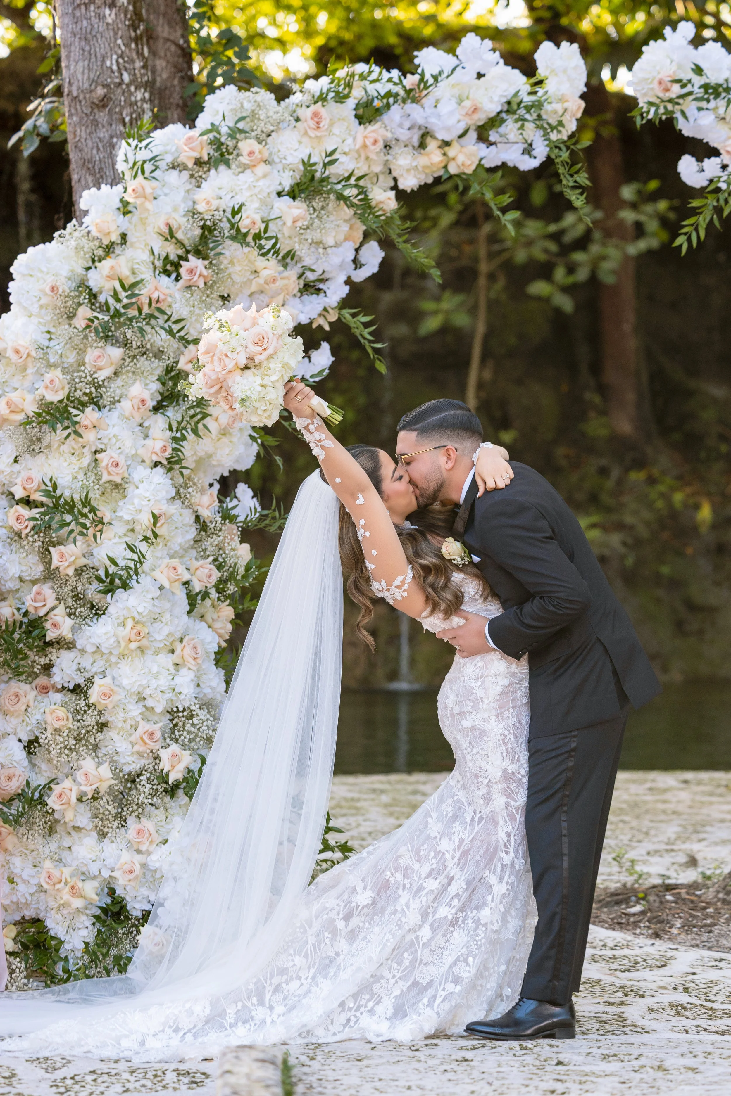 A newlywed couple sharing a kiss under a floral arch, with the bride raising a bouquet and wearing a lace wedding dress and long veil, and the groom in a black tuxedo, outdoors by a lake surrounded by trees.