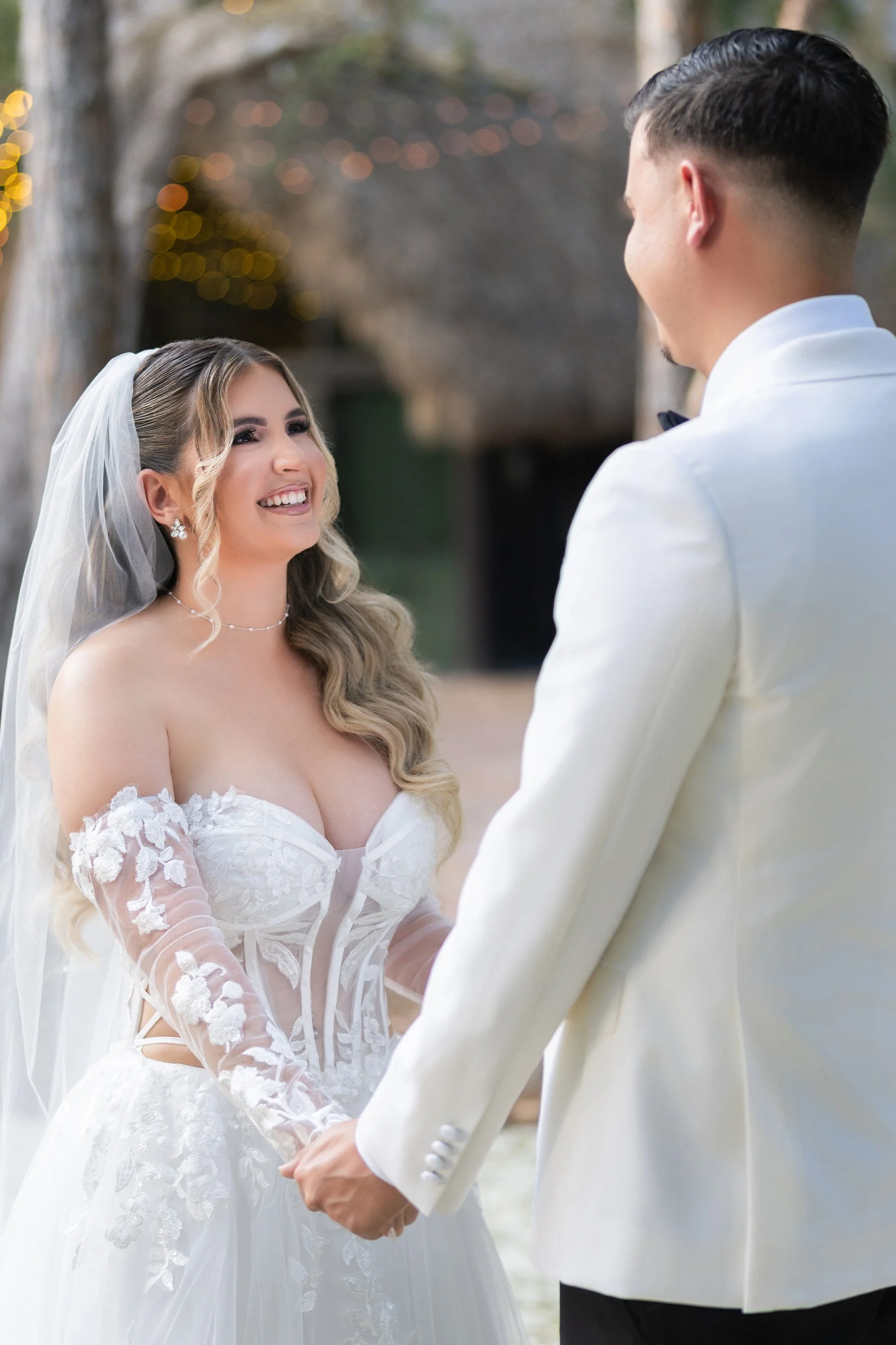 Bride and groom holding hands, standing outdoors during their wedding ceremony. The bride is smiling and wearing a white wedding dress with floral lace details, and the groom is dressed in a white tuxedo jacket.