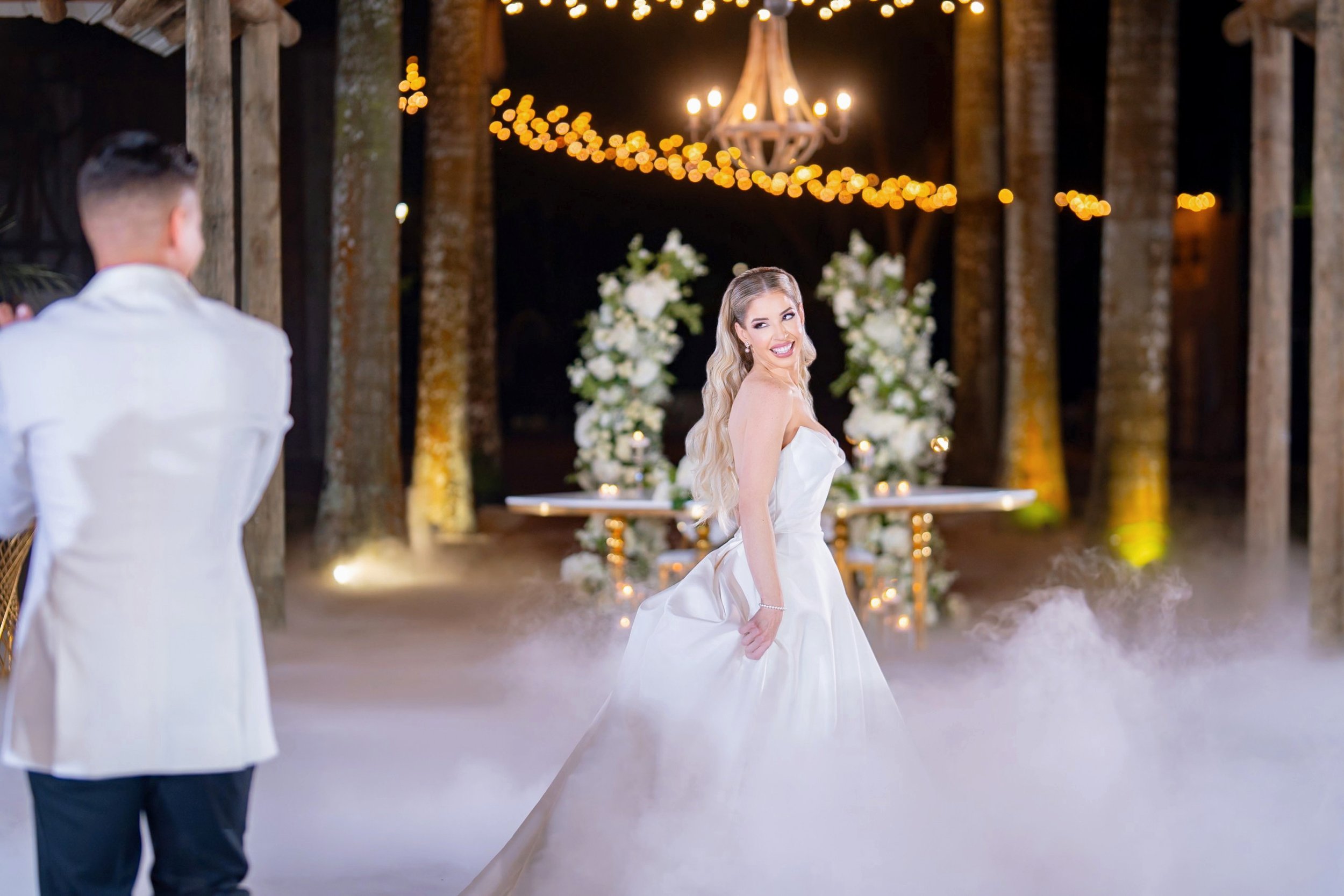 A bride in a white wedding dress smiling and holding her skirt, standing on a foggy dance floor with a groom in a white tuxedo facing her, at a wedding reception outdoor venue decorated with white flowers, candles, string lights, and a chandelier.