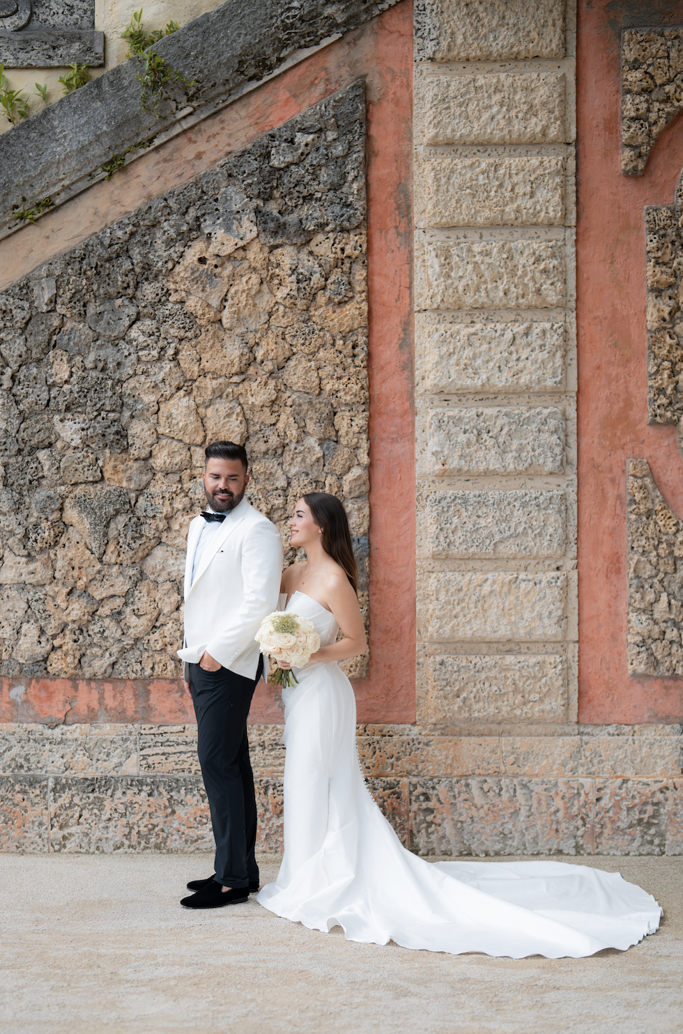 A bride and groom in wedding attire standing against a textured stone wall, the groom wearing a white tuxedo jacket and black pants, and the bride in a strapless white wedding gown holding a bouquet of white flowers.